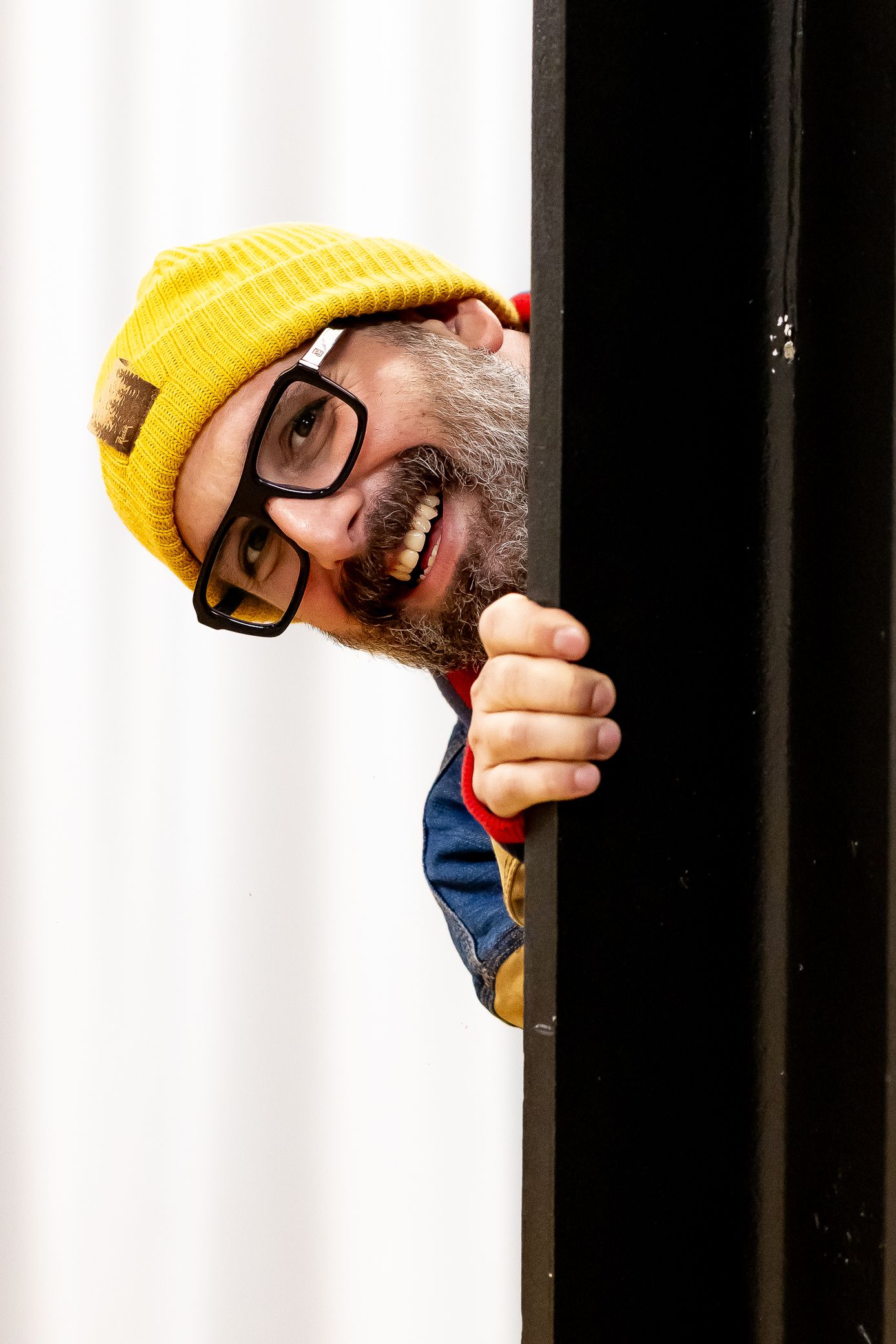 Funny bearded man in yellow beanie and glasses peeking around a black door frame with a big grin.