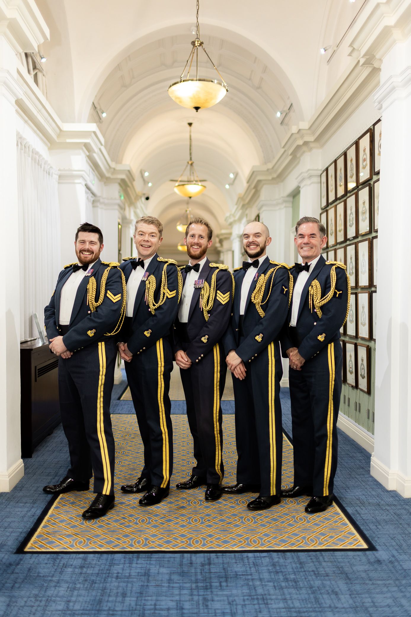 Five men in formal military dress uniforms with gold trim posing in an elegant hallway.
