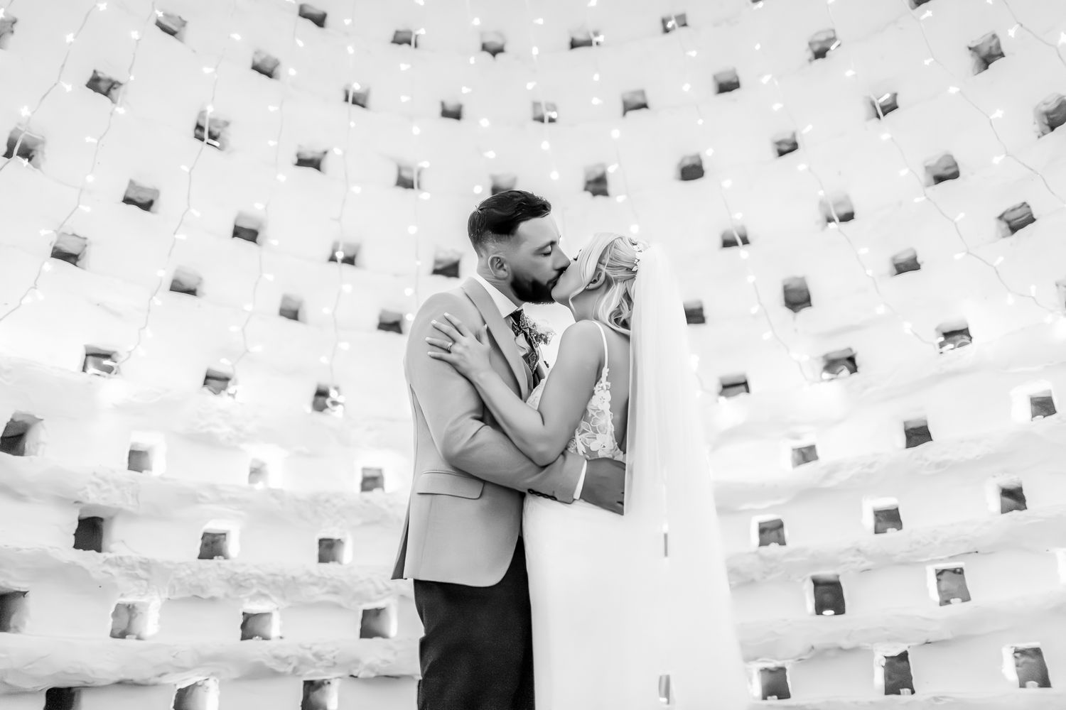 Black and white photo of a bride and groom embracing inside a decorative domed venue.