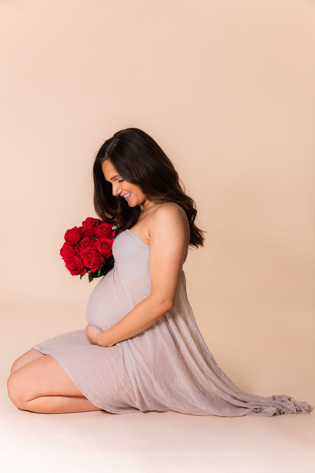 Smiling pregnant woman in a grey wrap dress holding red roses while sitting on studio floor.