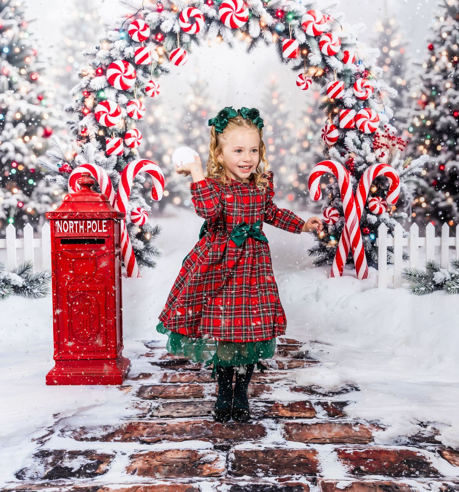 Girl in red plaid dress poses by North Pole mailbox under candy cane arch in snowy Christmas scene.