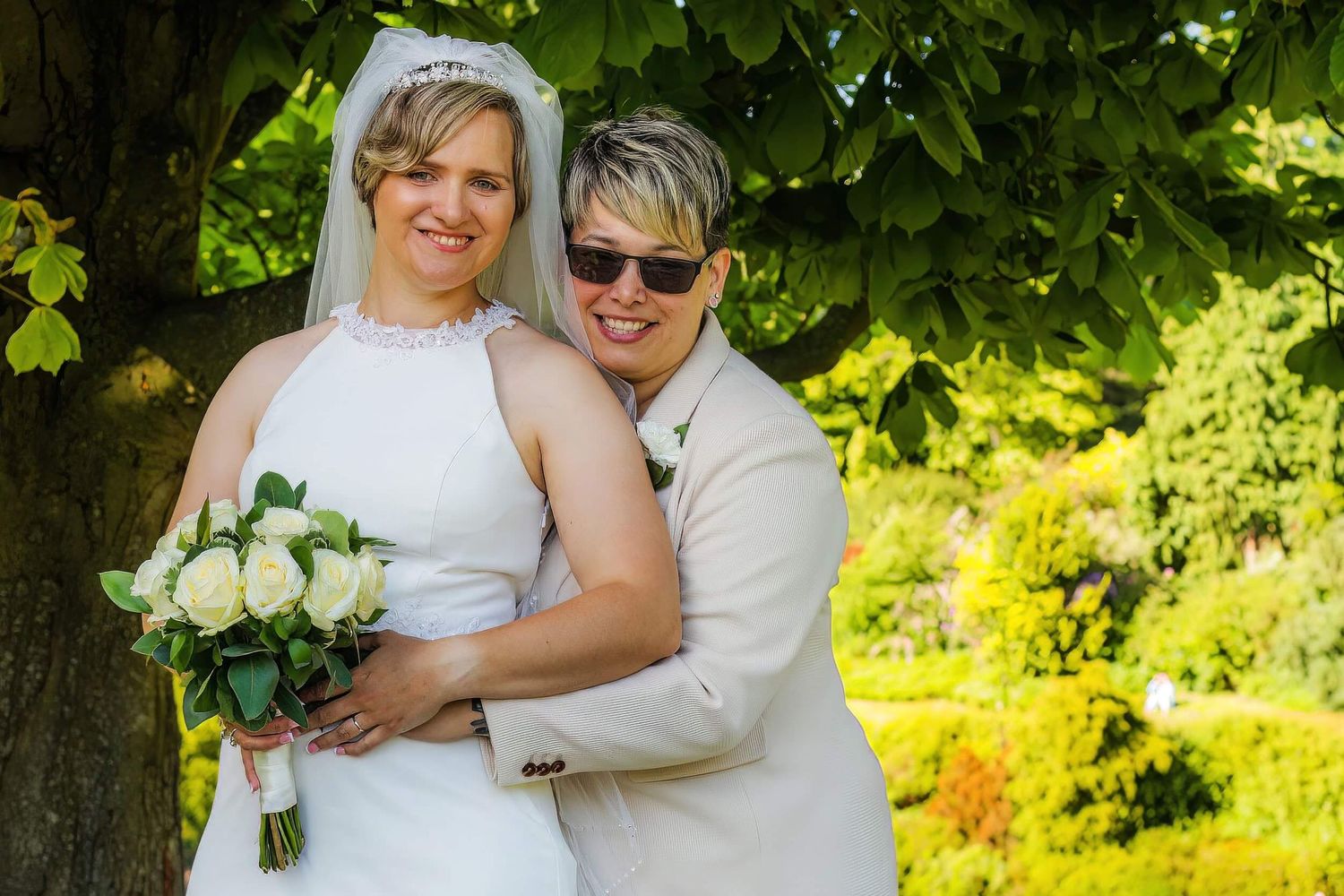 Two brides posing together under a tree, one in white gown holding bouquet, sunny garden wedding.