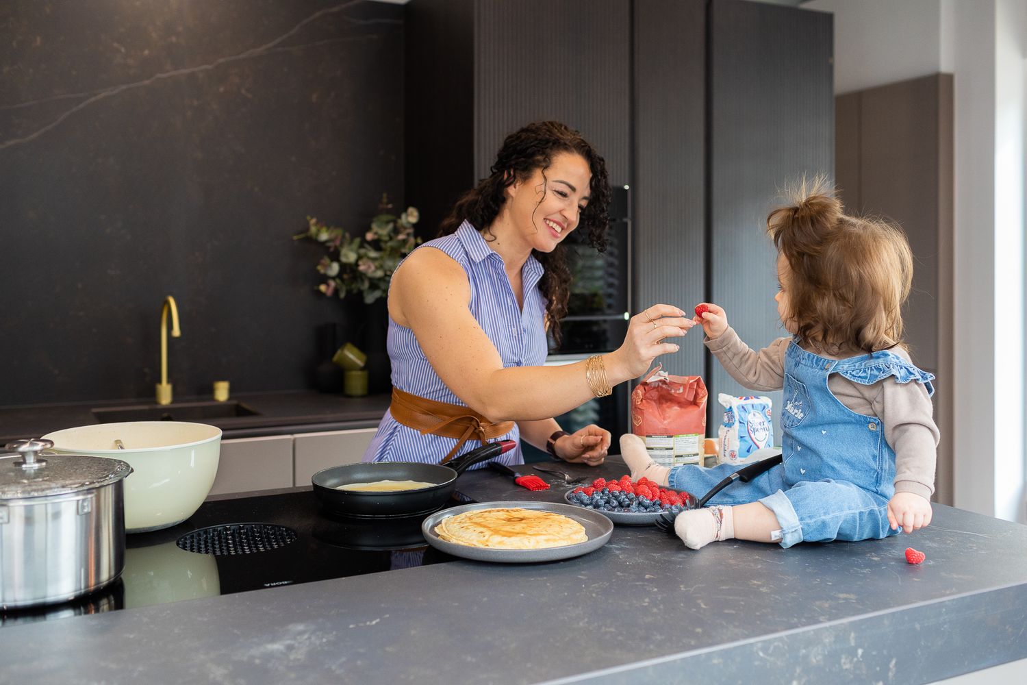 Happy mother and toddler cooking together in a modern dark kitchen with gold fixtures.