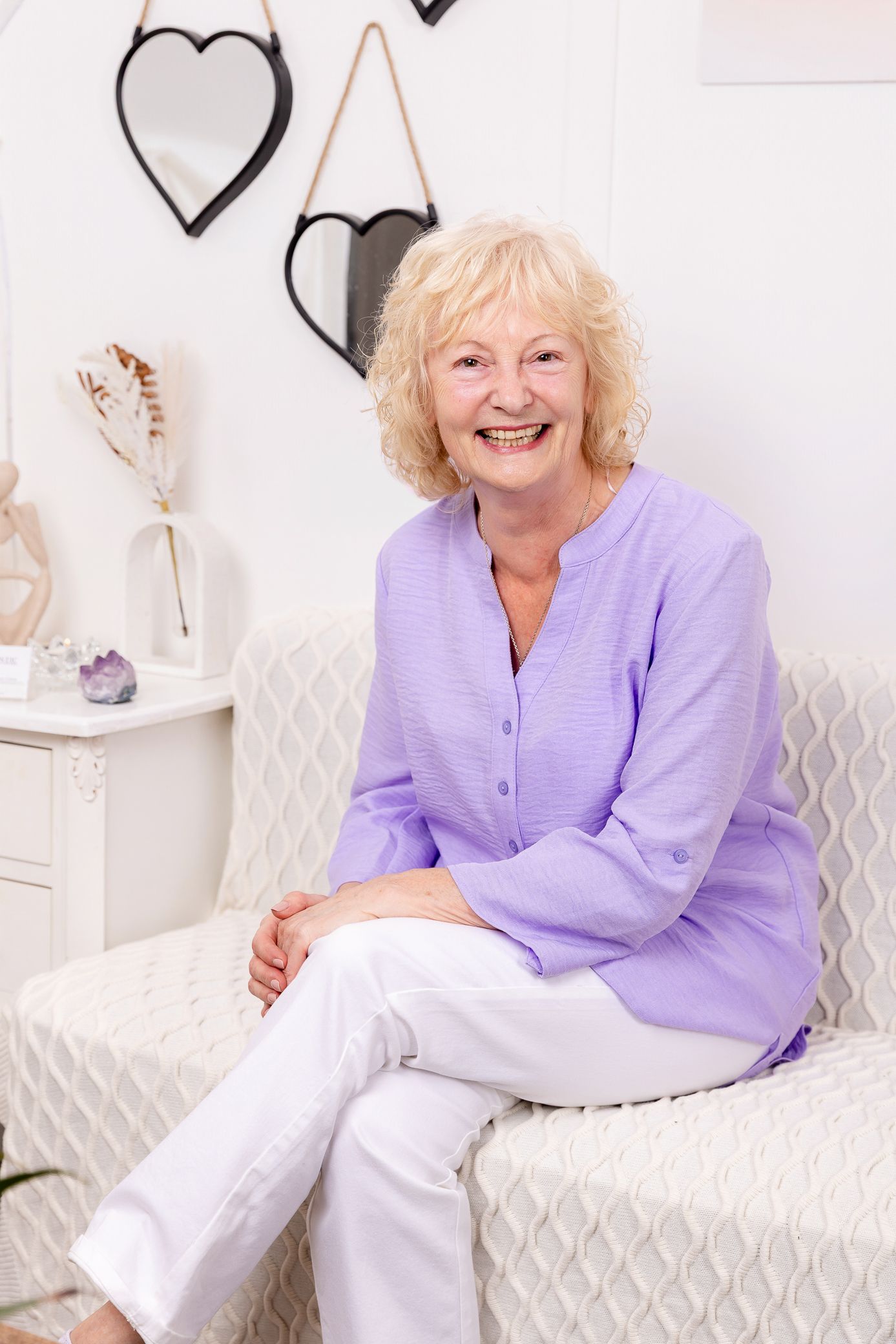Happy senior blonde woman in purple blouse sitting on a white chair smiling.