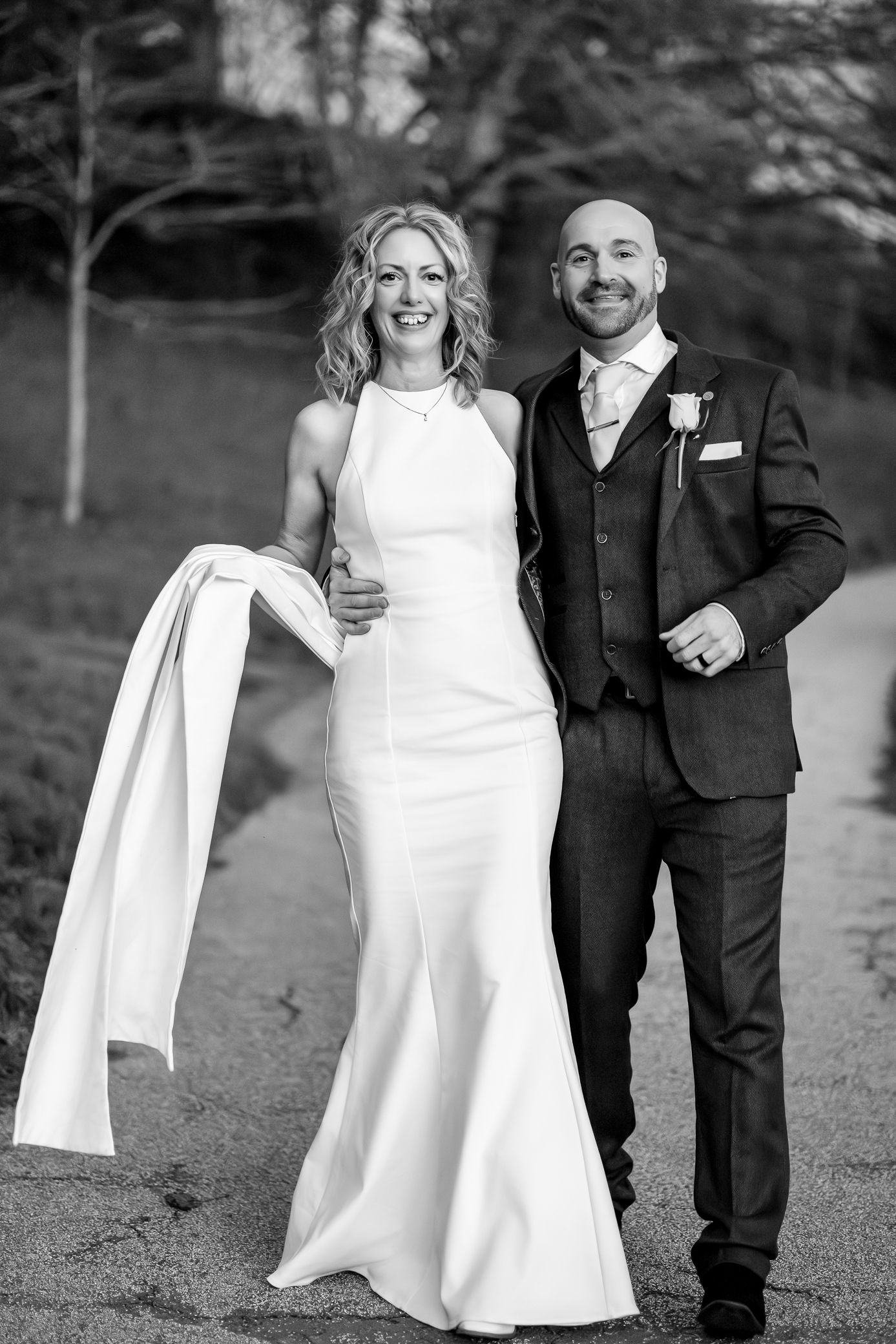 Black and white photo of a smiling bride in a white gown and groom in a suit walking together on their wedding day.