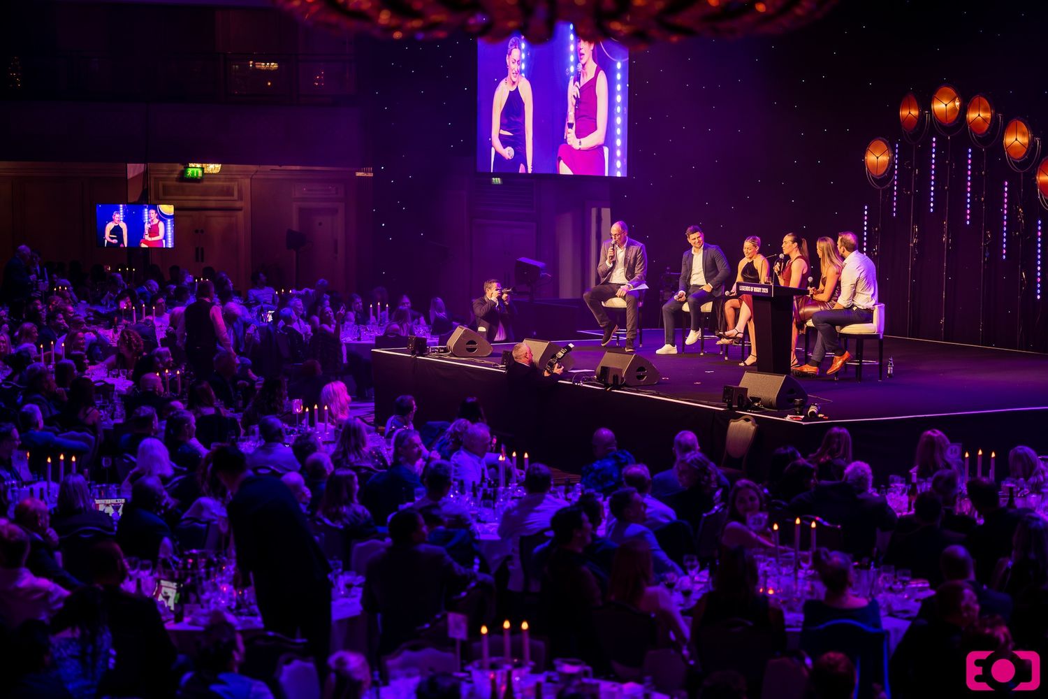 Elegant gala event with panelists on stage, large audience seated at tables under purple lighting in grand venue.