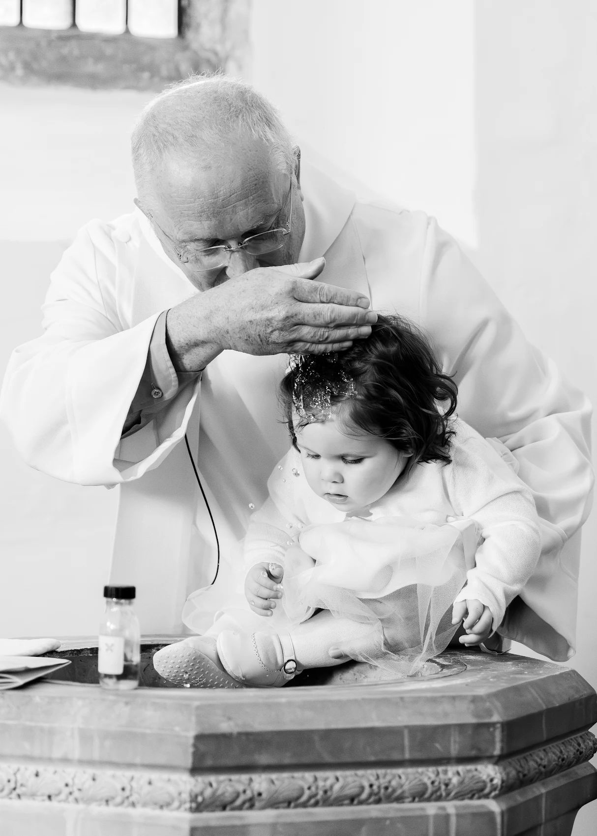 Black and white photo of a priest performing a baptism on a baby girl at a church font.