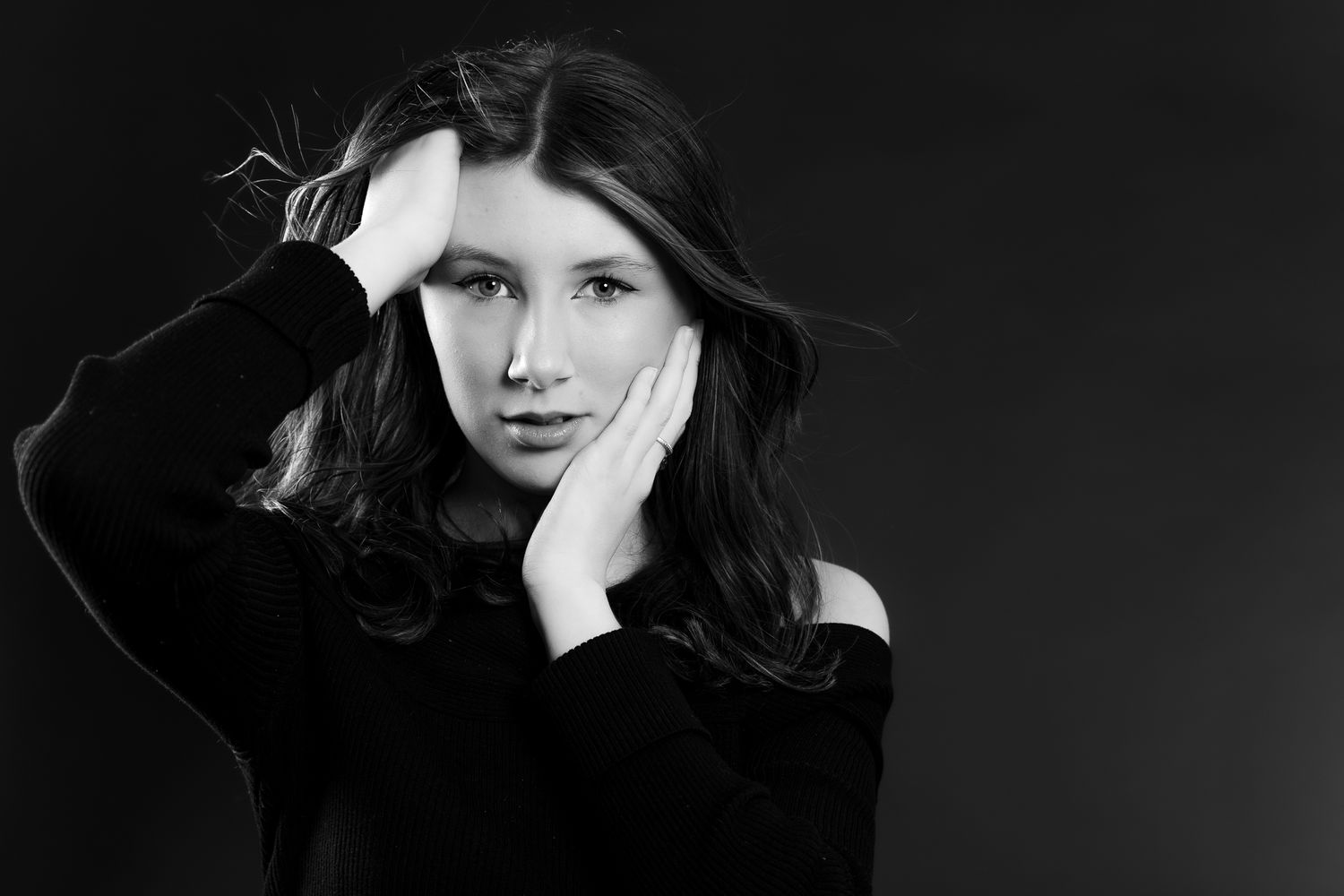 Black and white portrait of a young brunette woman in black top holding her face with both hands.