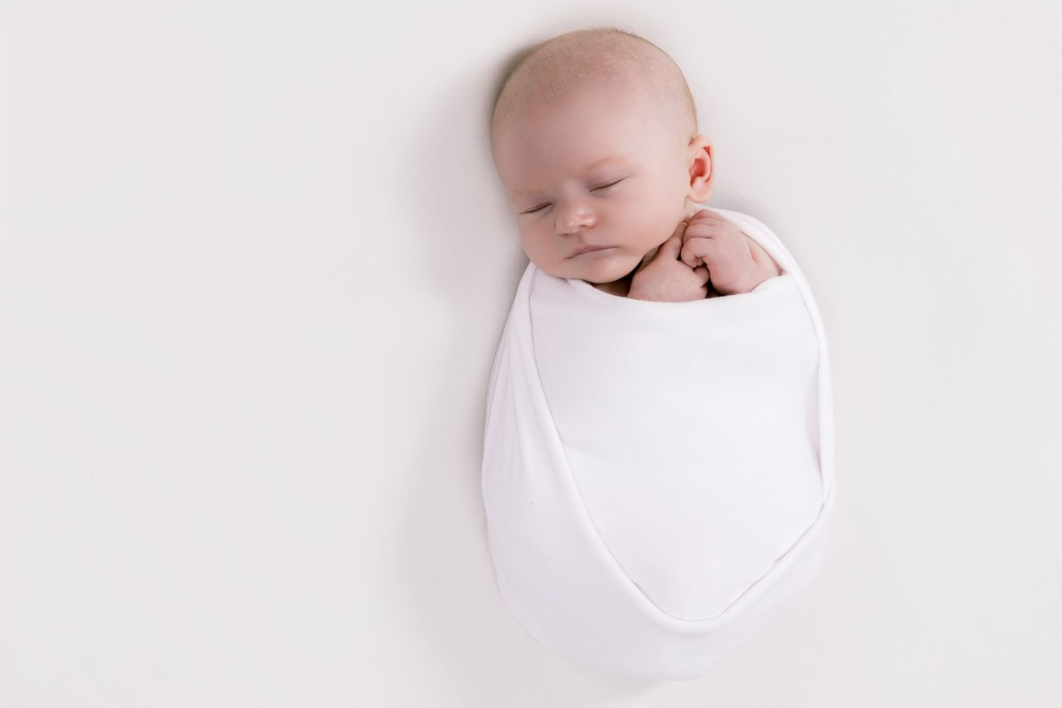 Peaceful newborn baby sleeping soundly wrapped in a white swaddle on a white background.