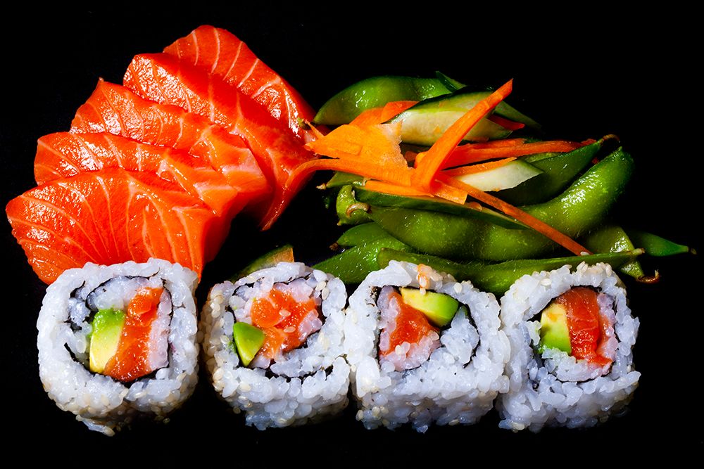 Salmon sashimi, California rolls, edamame, and pickled vegetables on a black background.
