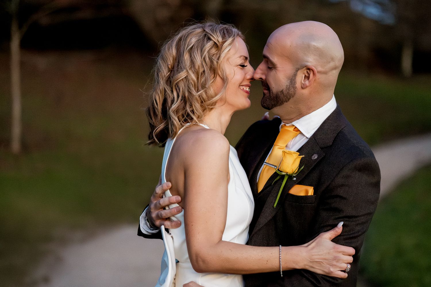 Bride and bald groom share romantic moment outdoors, groom wearing yellow tie and boutonniere.