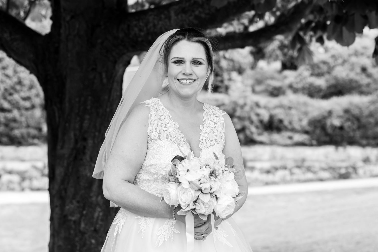 Bride in lace wedding dress and veil holding bouquet, smiling outdoors under tree in black and white photo.
