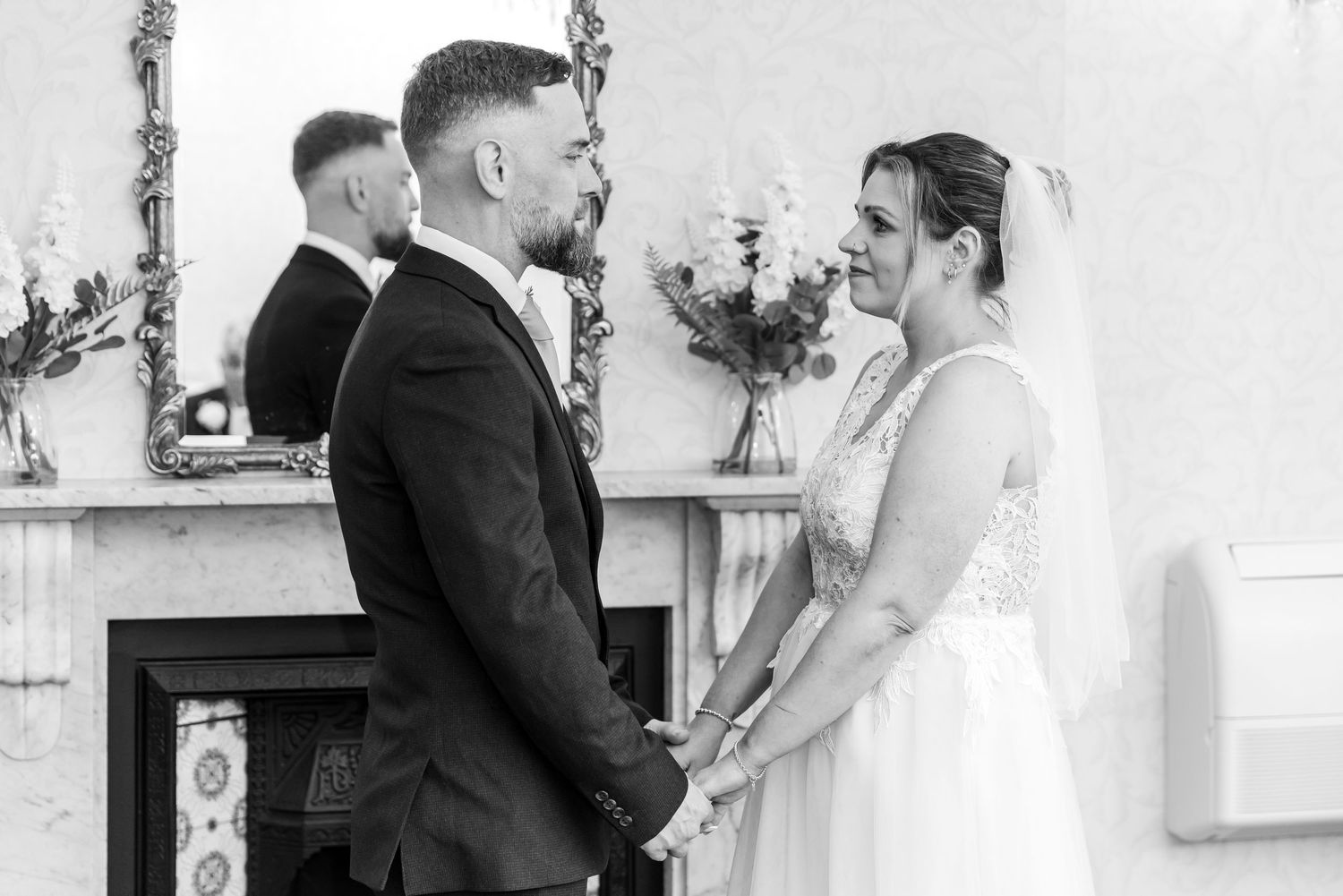 Black and white wedding photo of bride and groom holding hands at altar before fireplace with floral arrangement.