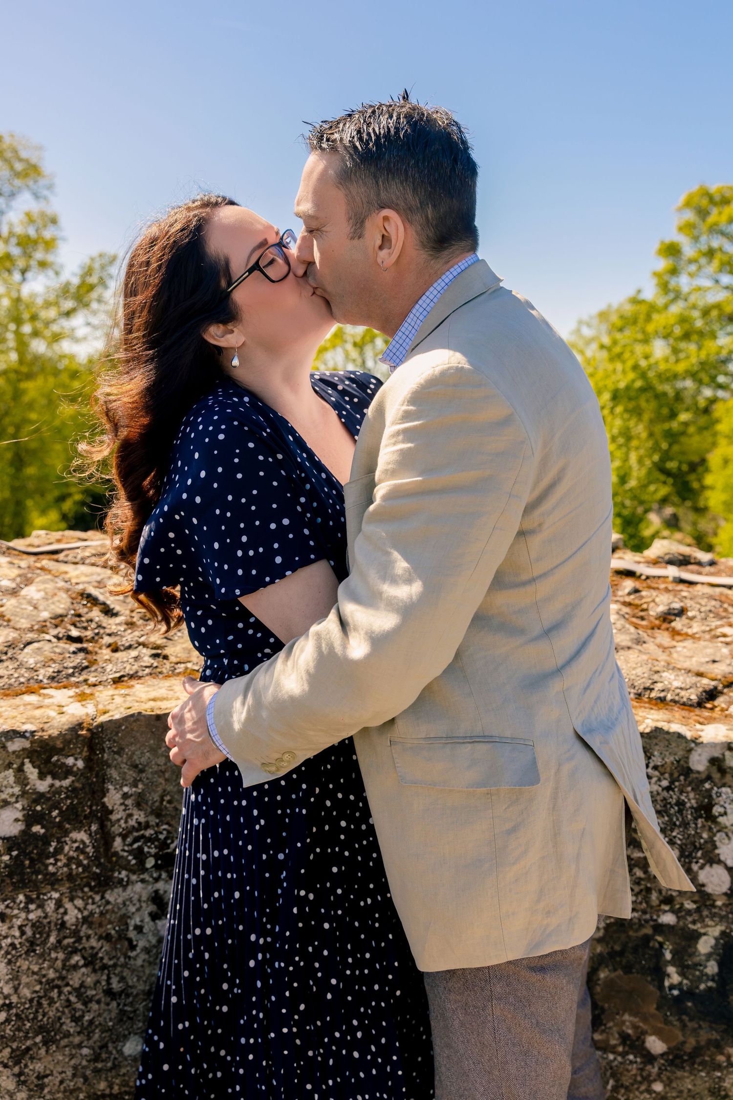 Couple sharing a romantic kiss outdoors near a stone wall, woman in navy polka dot dress and man in beige blazer.