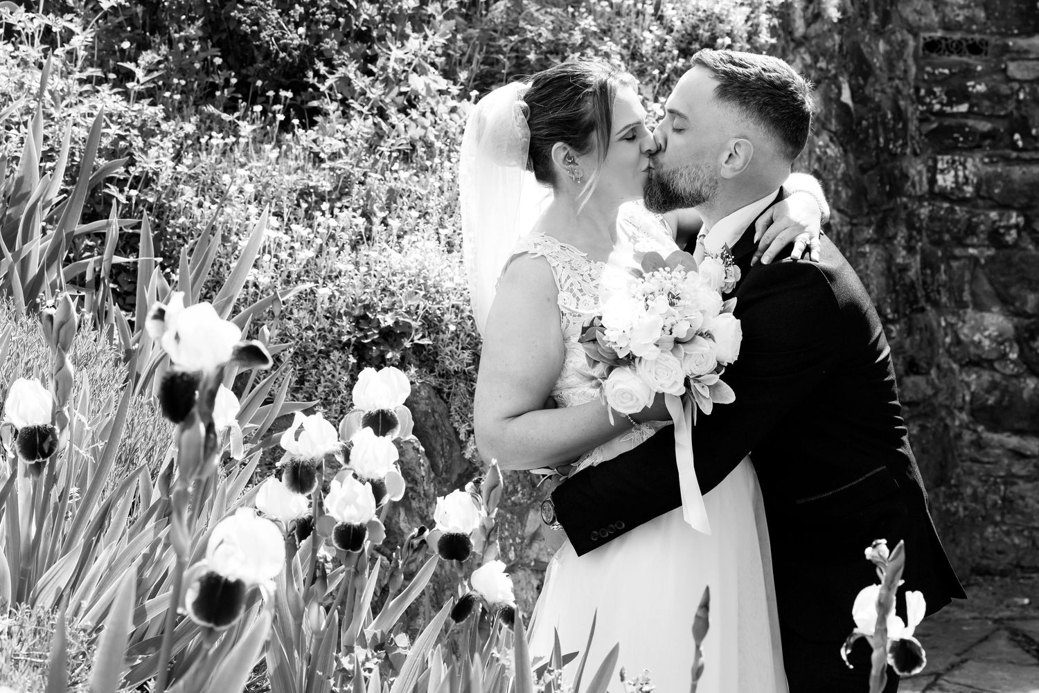Black and white photo of a bride and groom kissing among iris flowers in a garden on their wedding day.