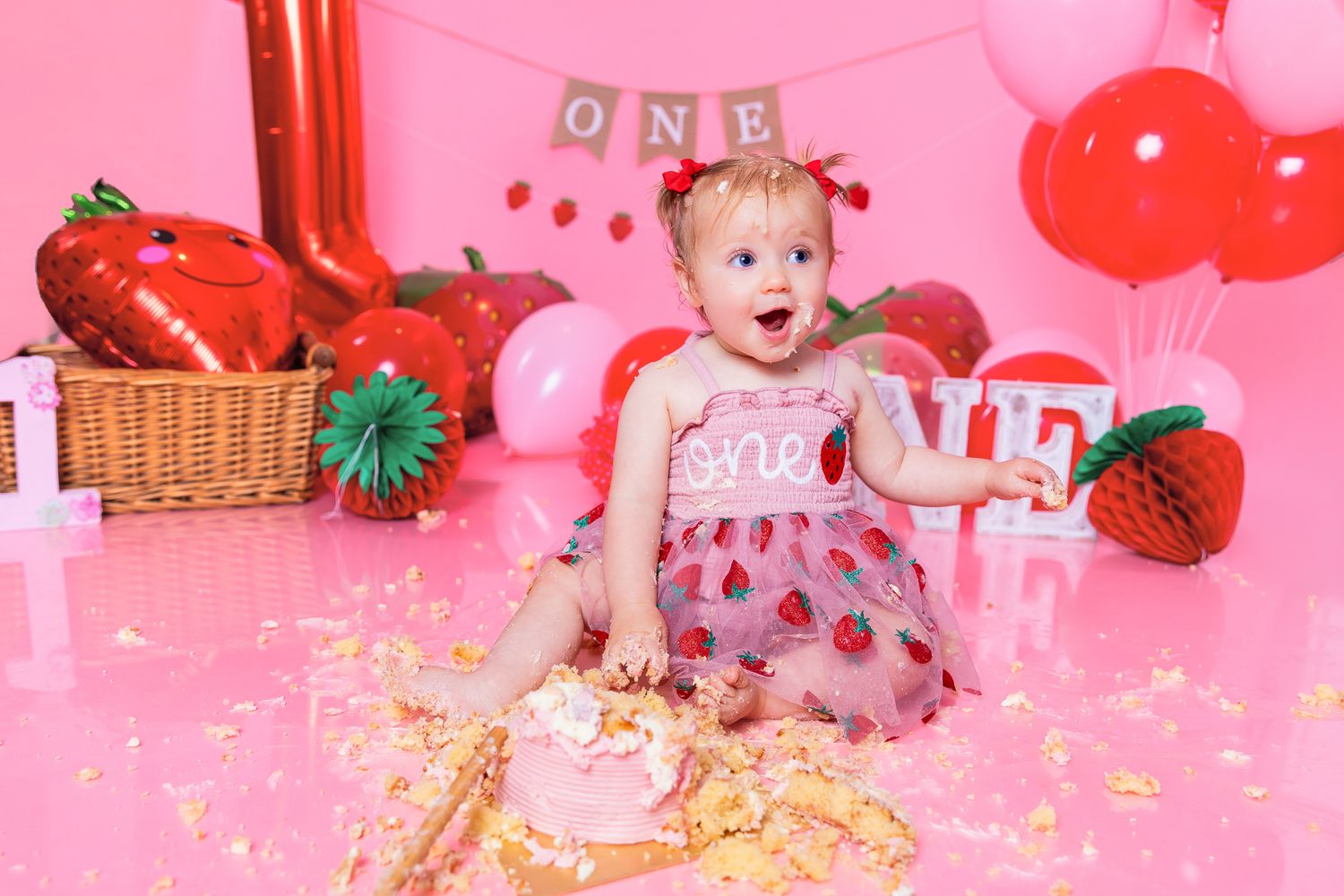 Baby girl in strawberry dress celebrates first birthday smashing cake on pink backdrop with ONE banner and balloons.