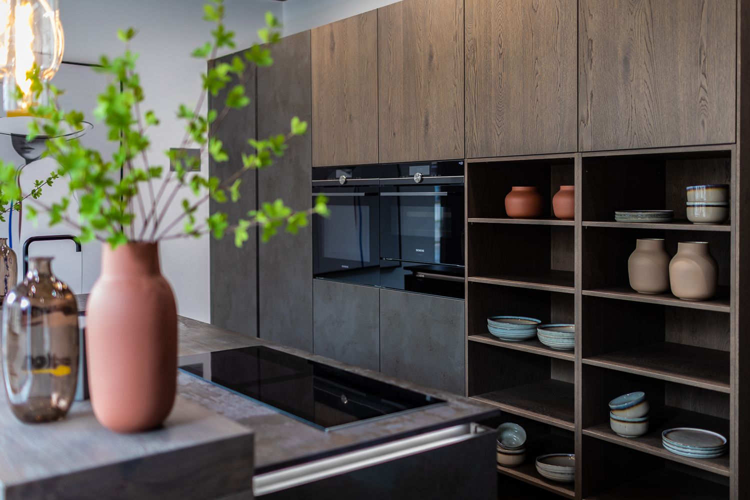 Modern kitchen with wood-effect cabinetry, built-in oven, open shelves displaying colorful ceramic bowls and vases.