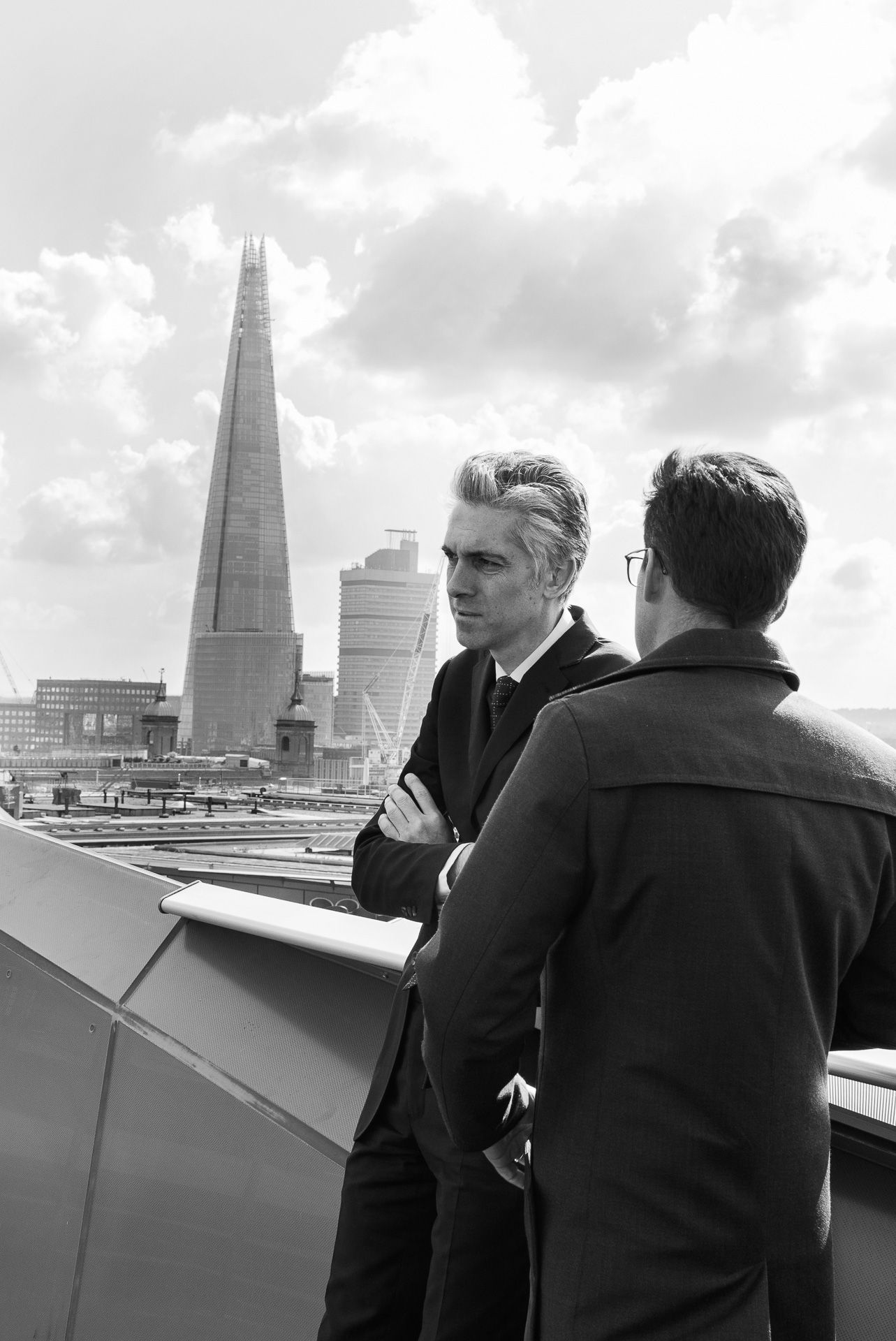 Two men in suits conversing on a rooftop with The Shard skyscraper in the London skyline.