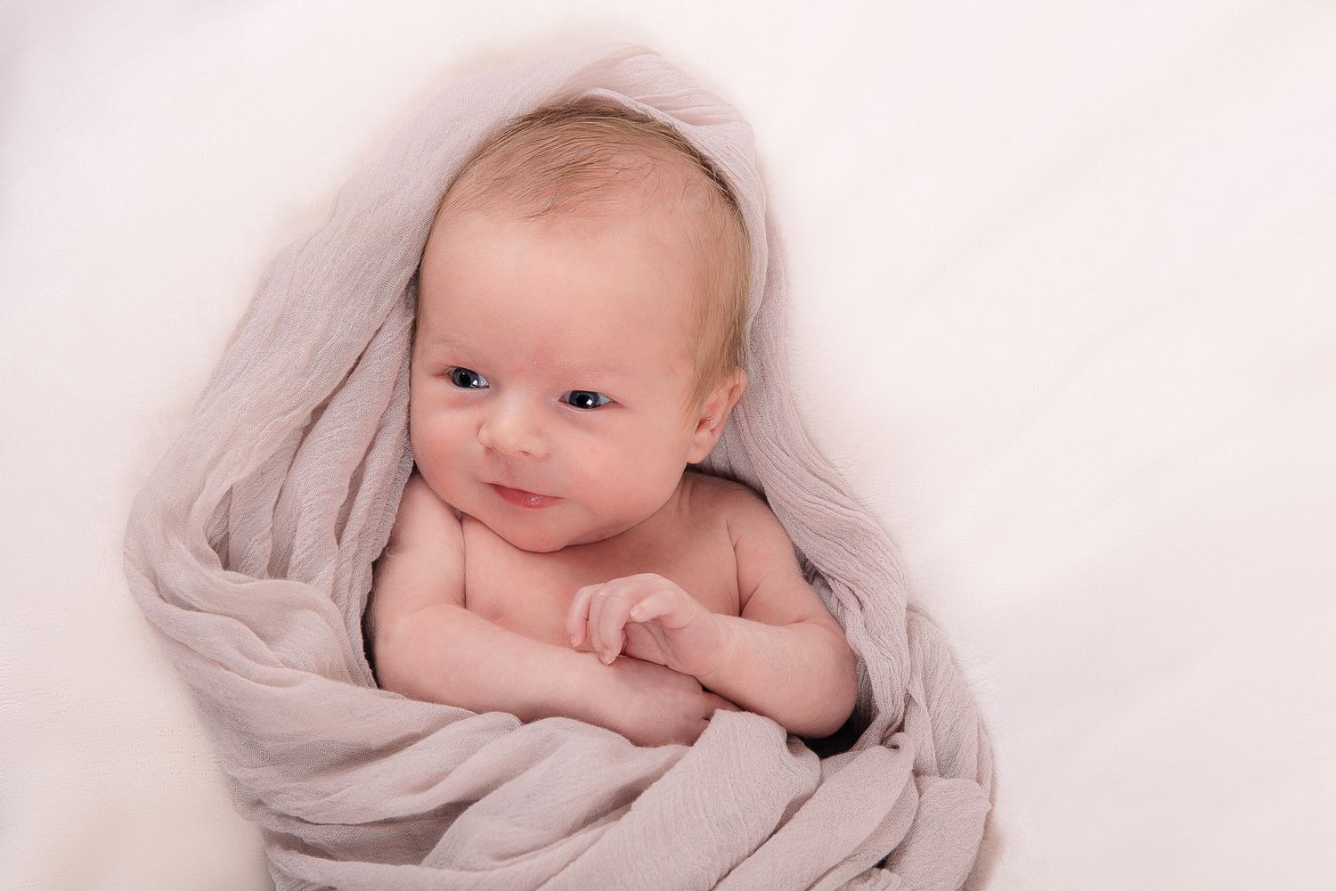Adorable newborn baby wrapped in a soft grey blanket smiling gently at the camera in a studio portrait.