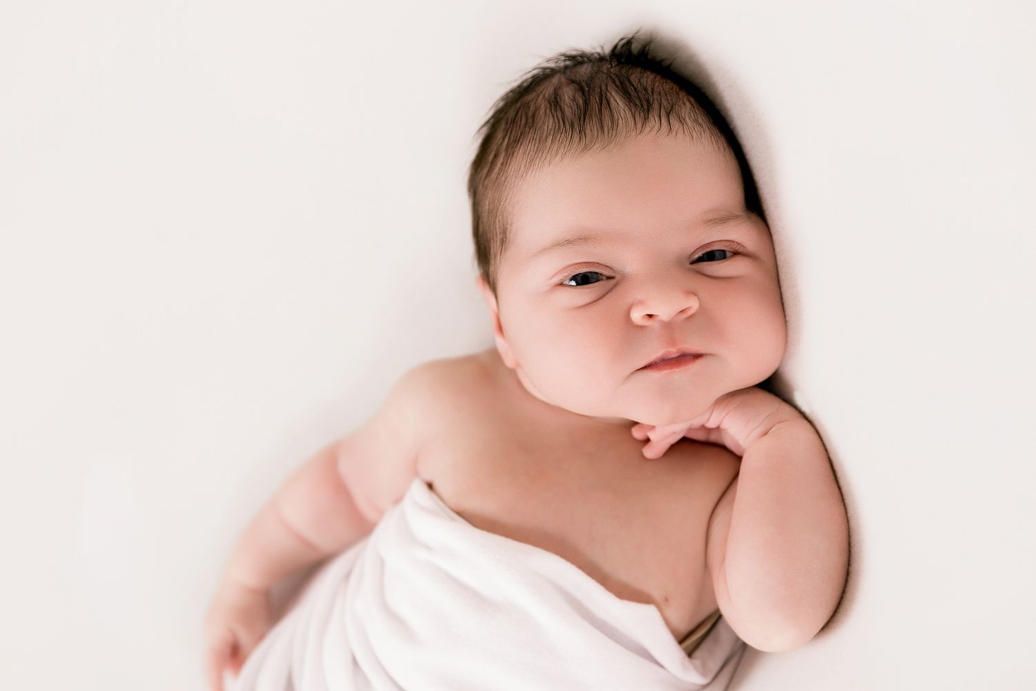 Alert newborn baby with dark hair resting chin on hand wrapped in white.