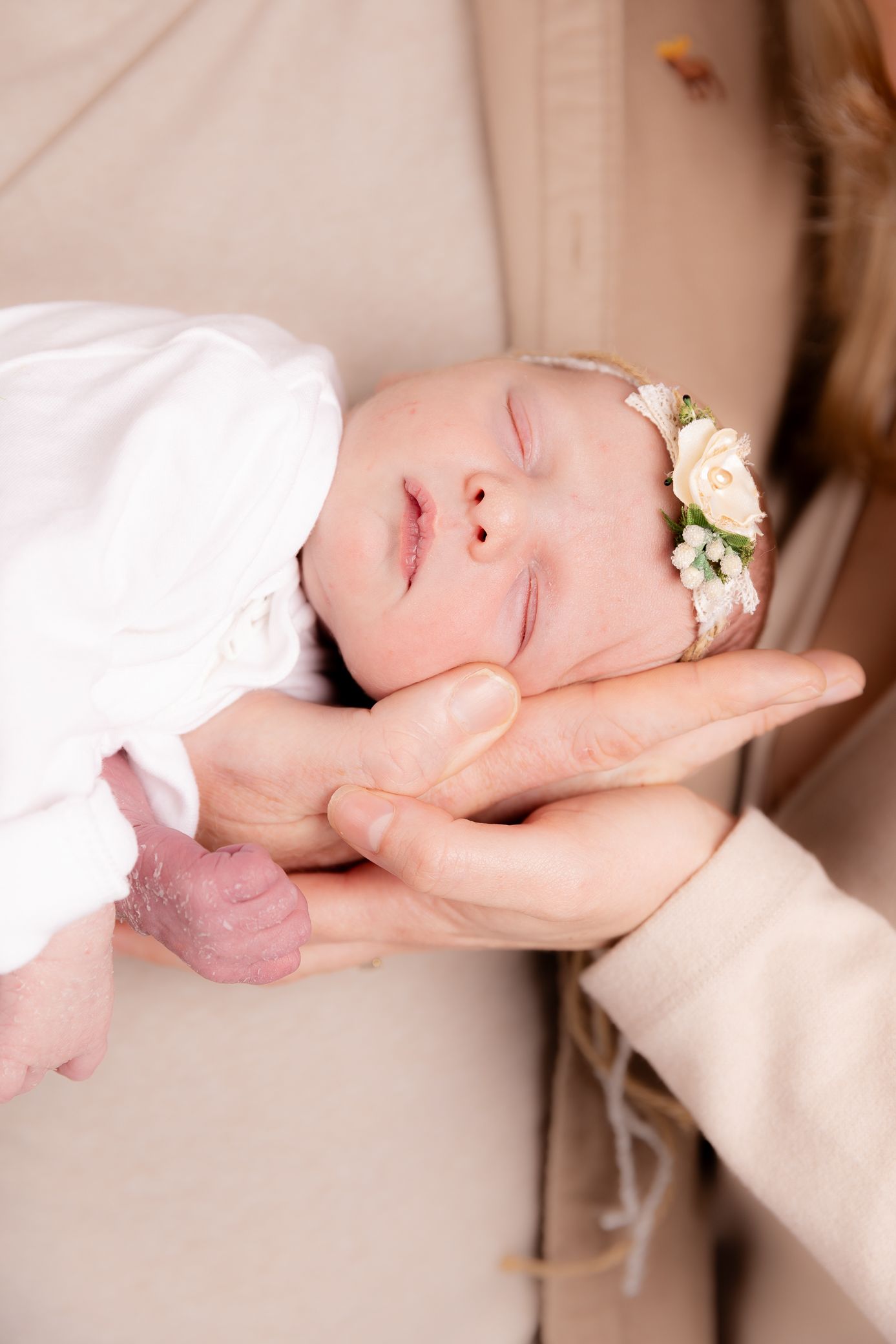Close-up of a sleeping newborn baby girl wearing a floral headband, cradled gently in a parents hands.