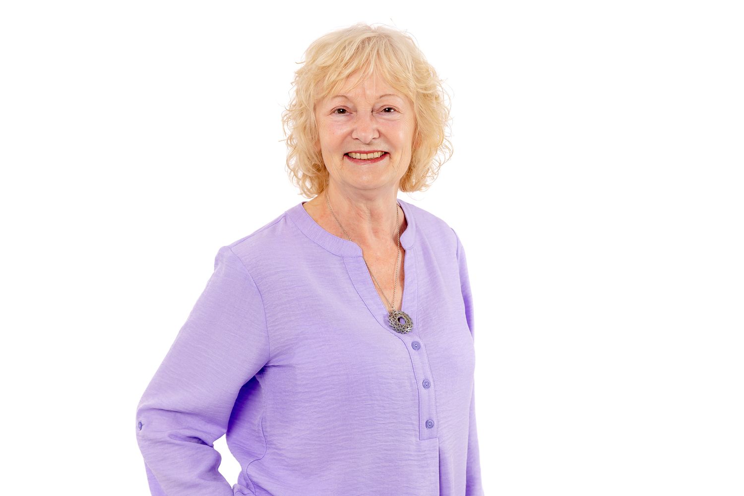 Smiling senior woman with blonde hair wearing a purple cardigan posing against a white background.