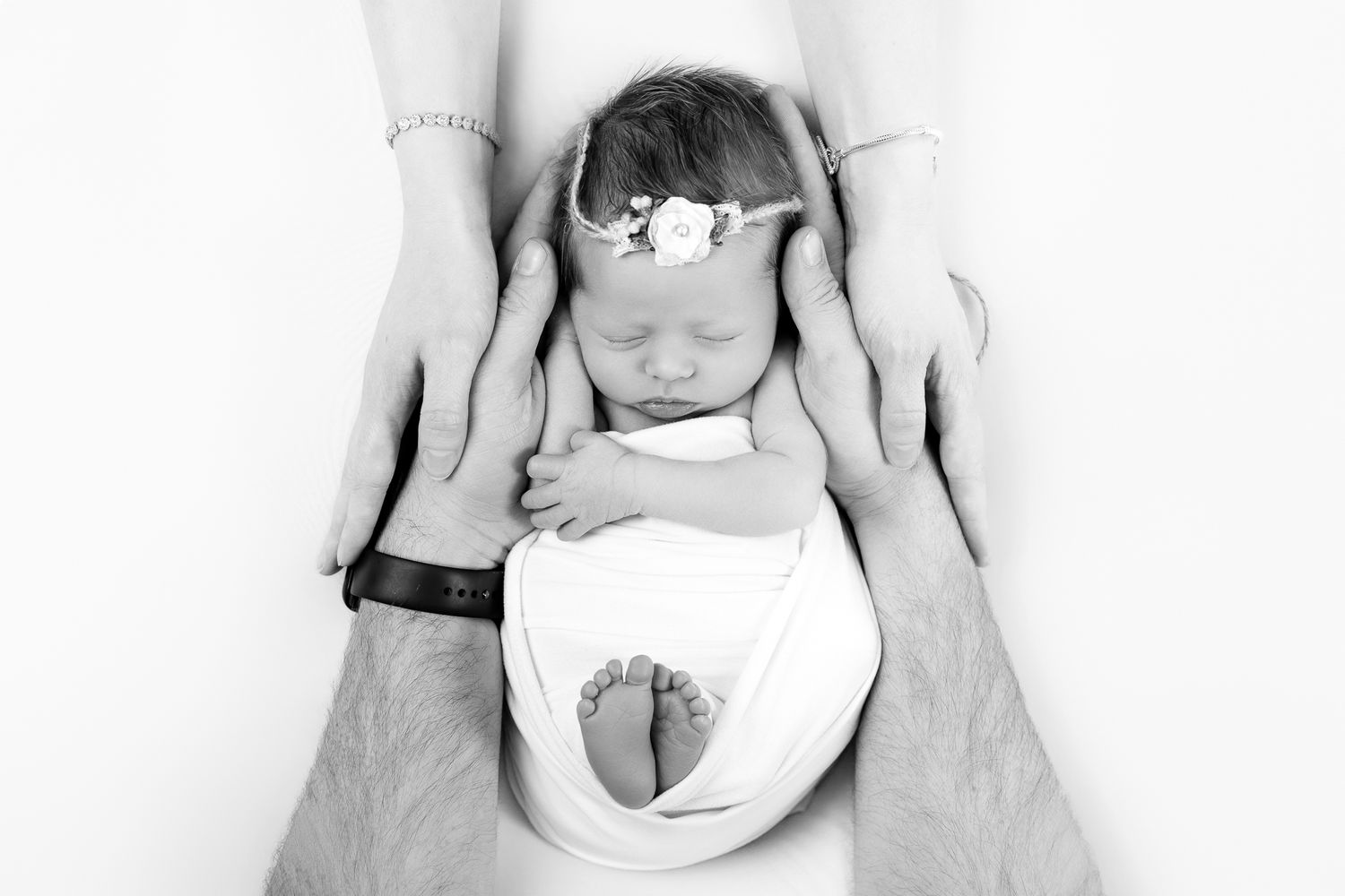 Black and white newborn photo of sleeping baby cradled by parents hands, wearing white wrap and floral headband.