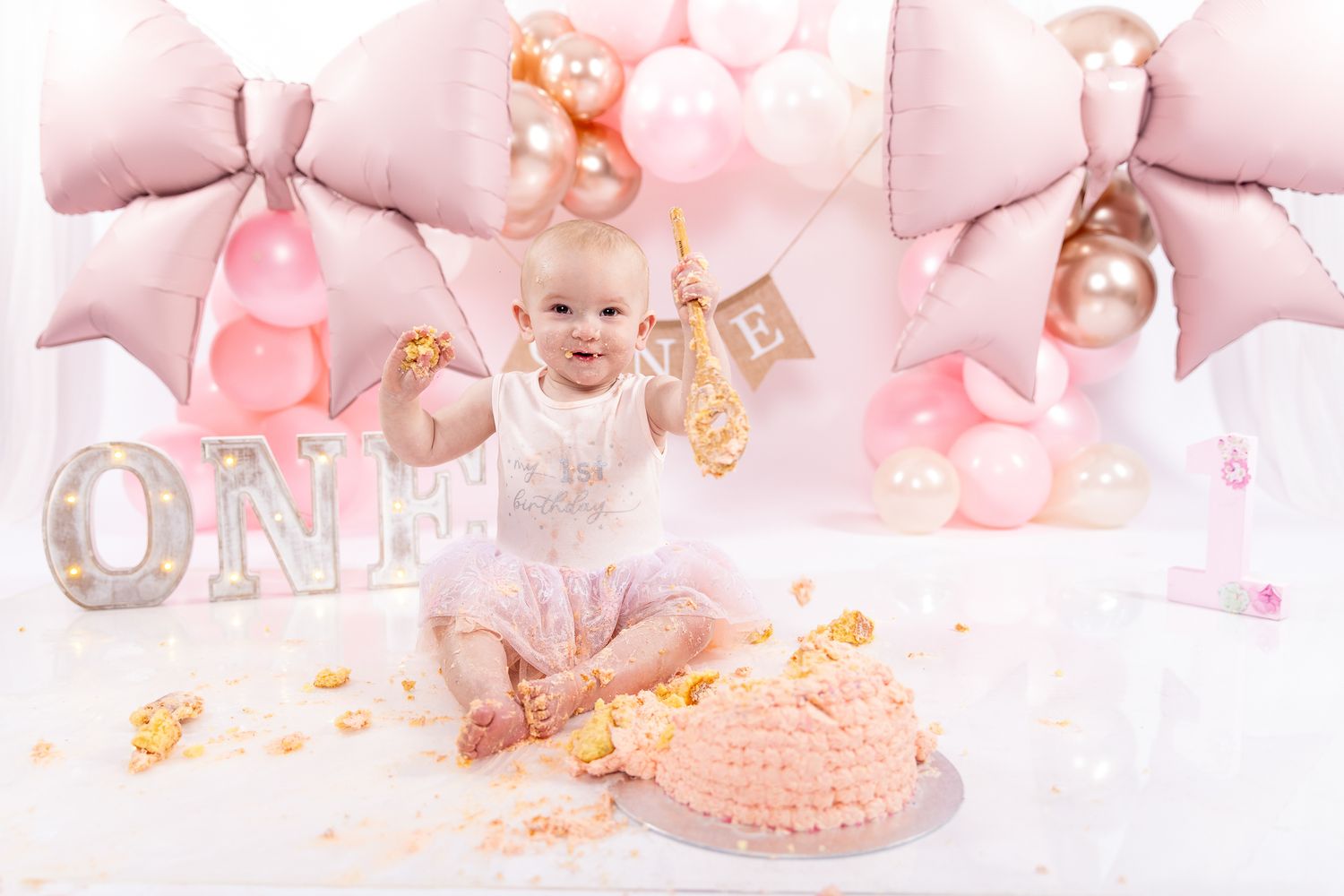 Baby girl celebrates first birthday smashing pink cake surrounded by balloons and ONE sign.