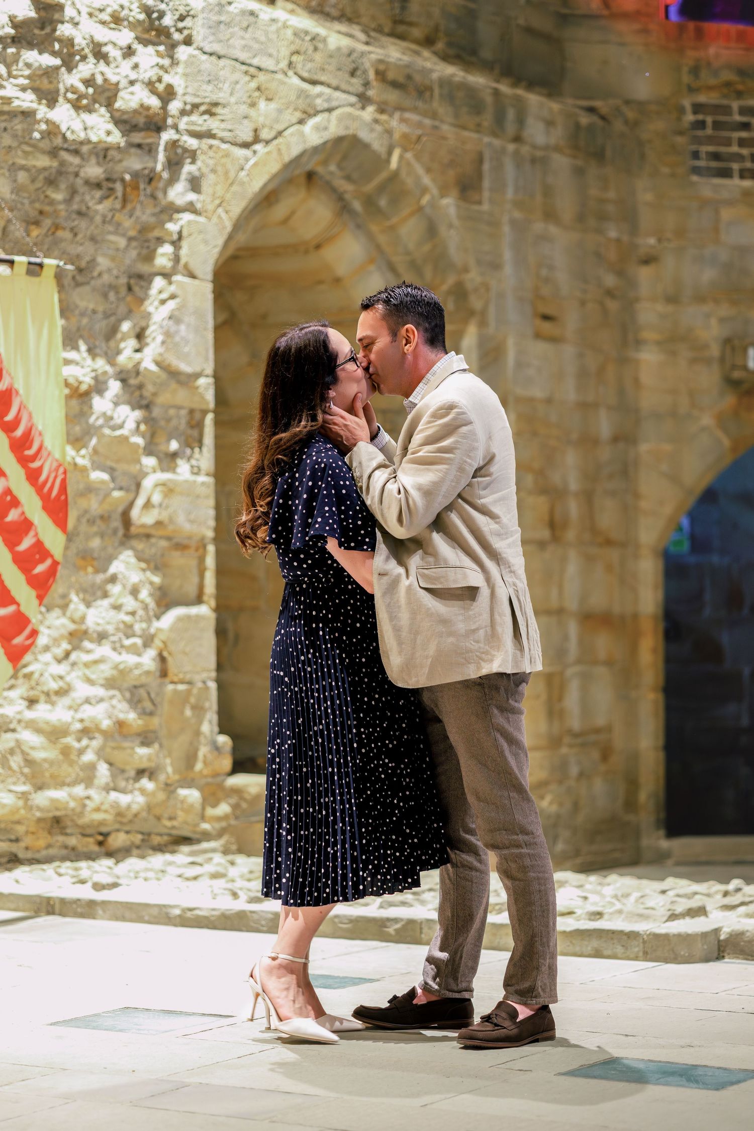 Couple sharing a romantic kiss in front of a historic stone archway, woman in navy dress, man in beige blazer.