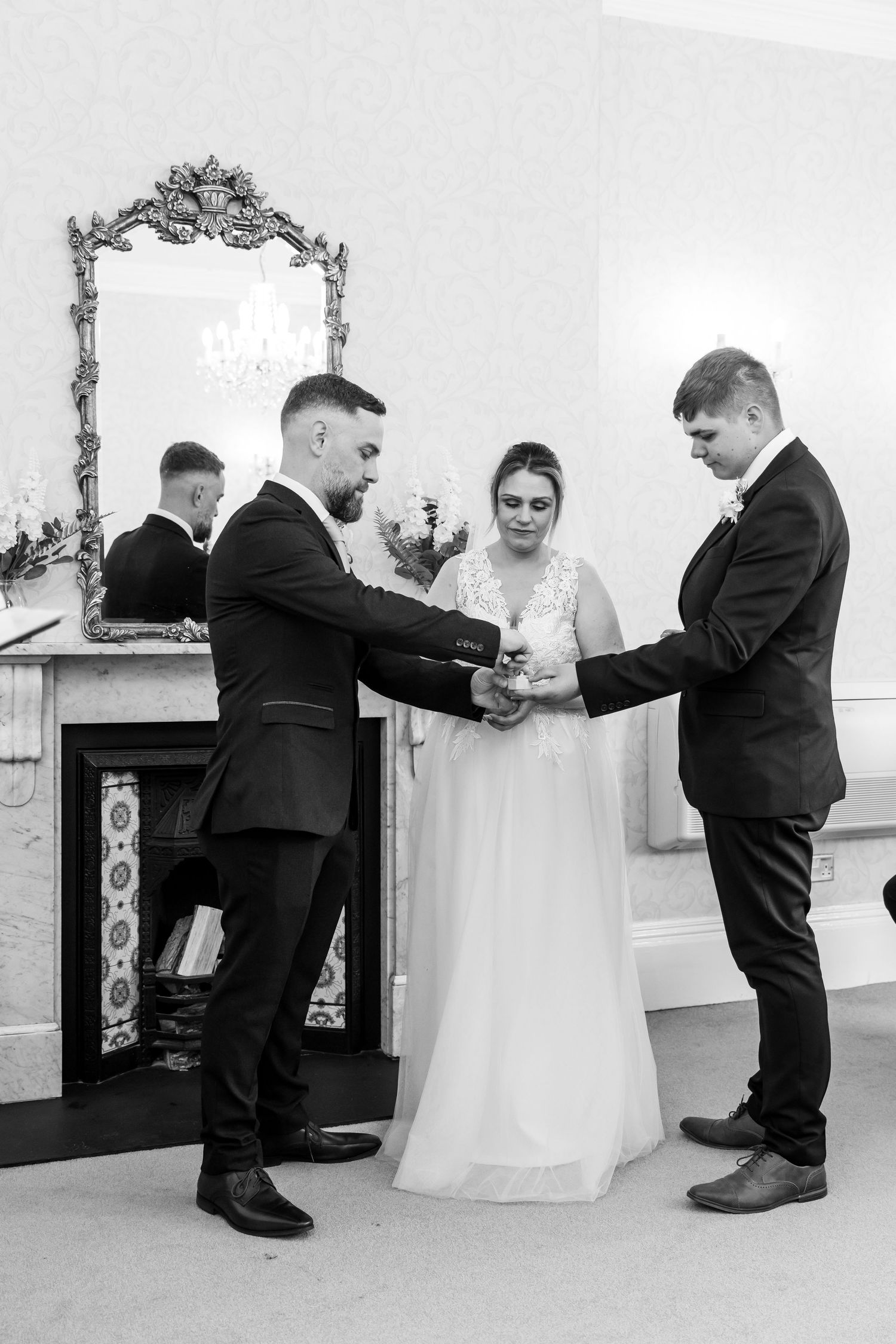 Black and white wedding ceremony photo of bride and groom exchanging rings with officiant by ornate fireplace.