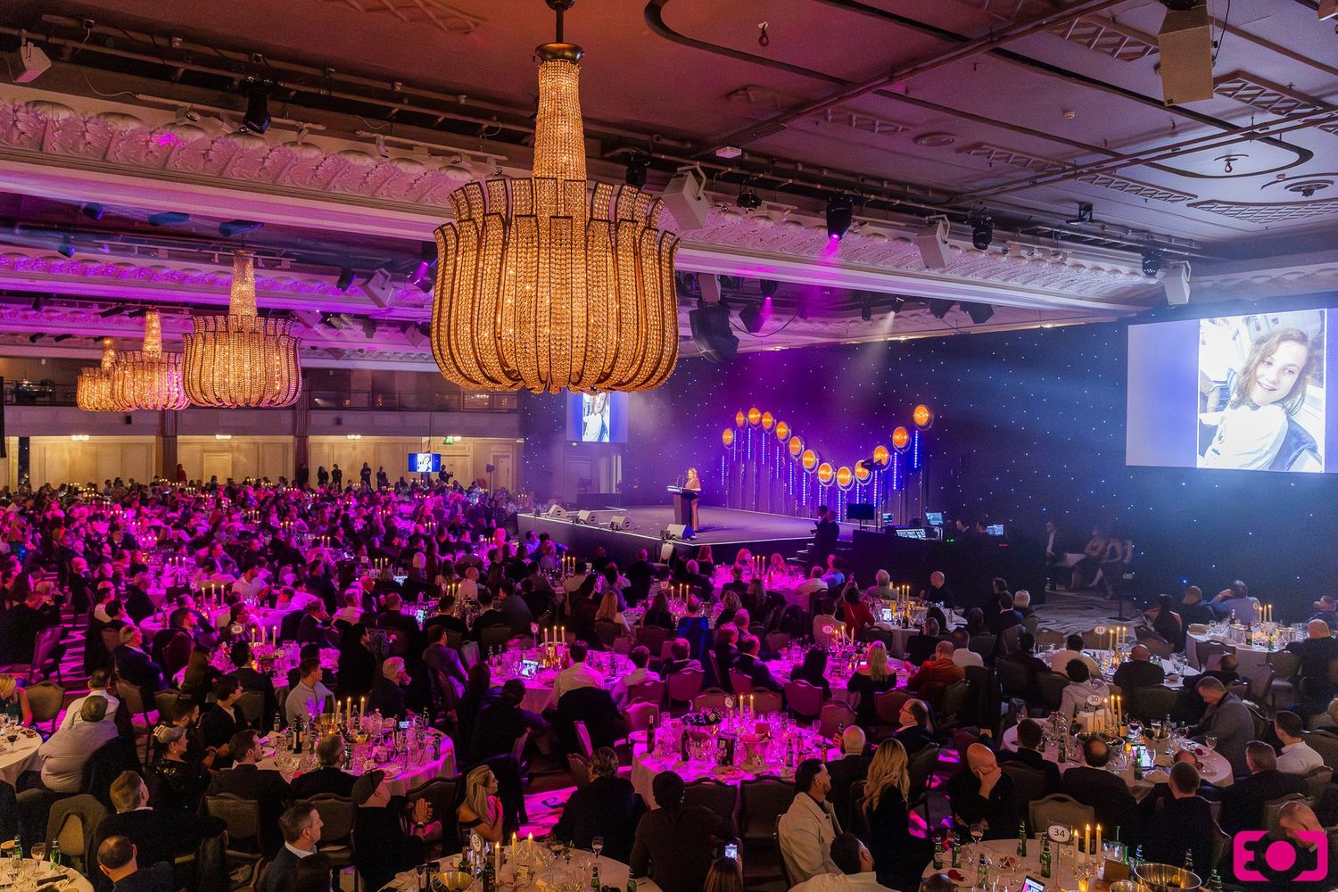 Elegant gala dinner event with chandeliers, purple lighting, packed round tables, and a large presentation screen on stage.