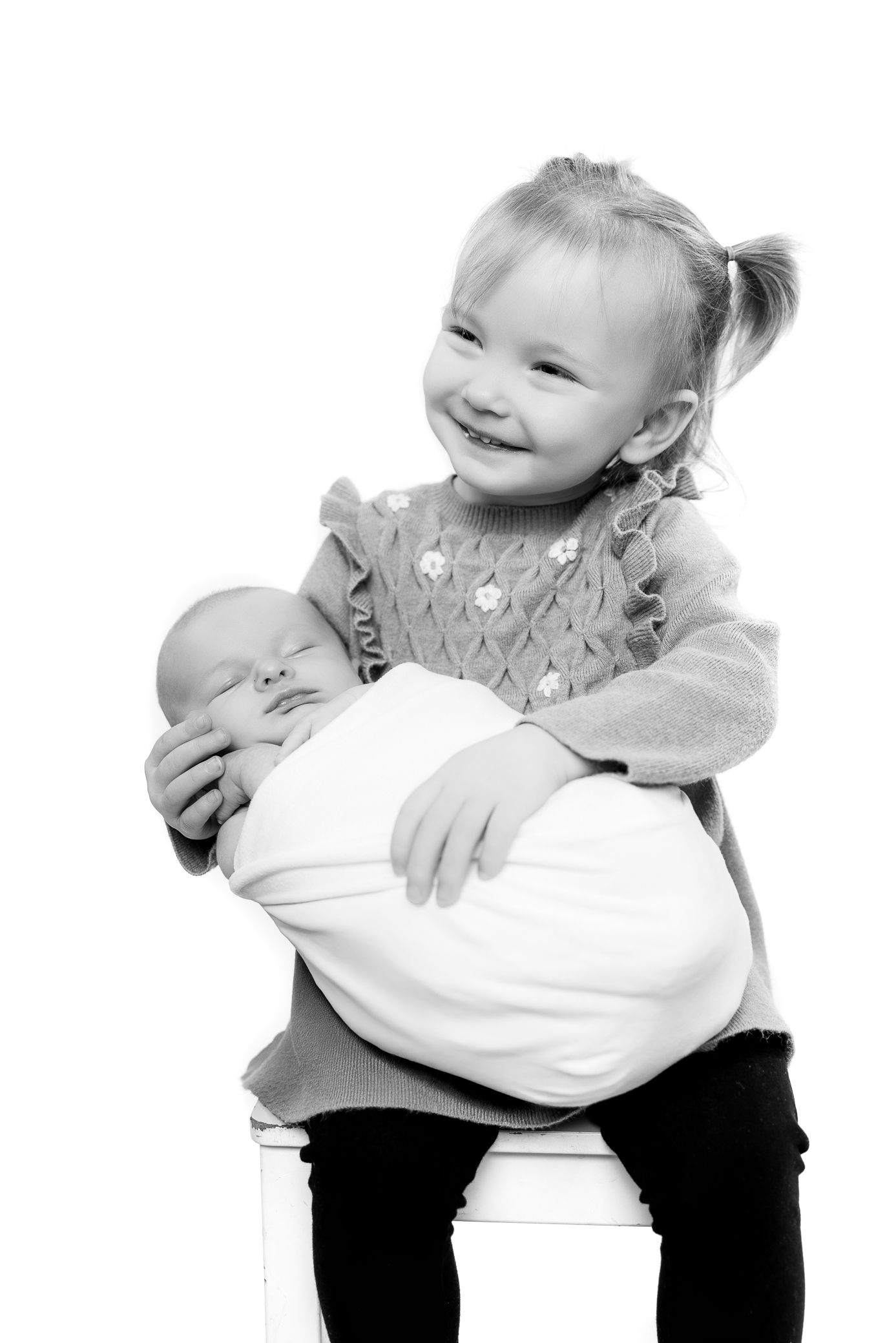 Smiling toddler girl holding a swaddled newborn baby sibling in a black and white portrait photo.