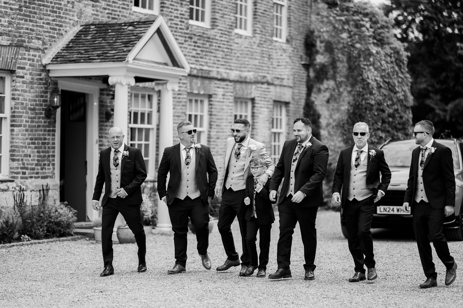 Black and white photo of groom and groomsmen walking confidently outside elegant manor.