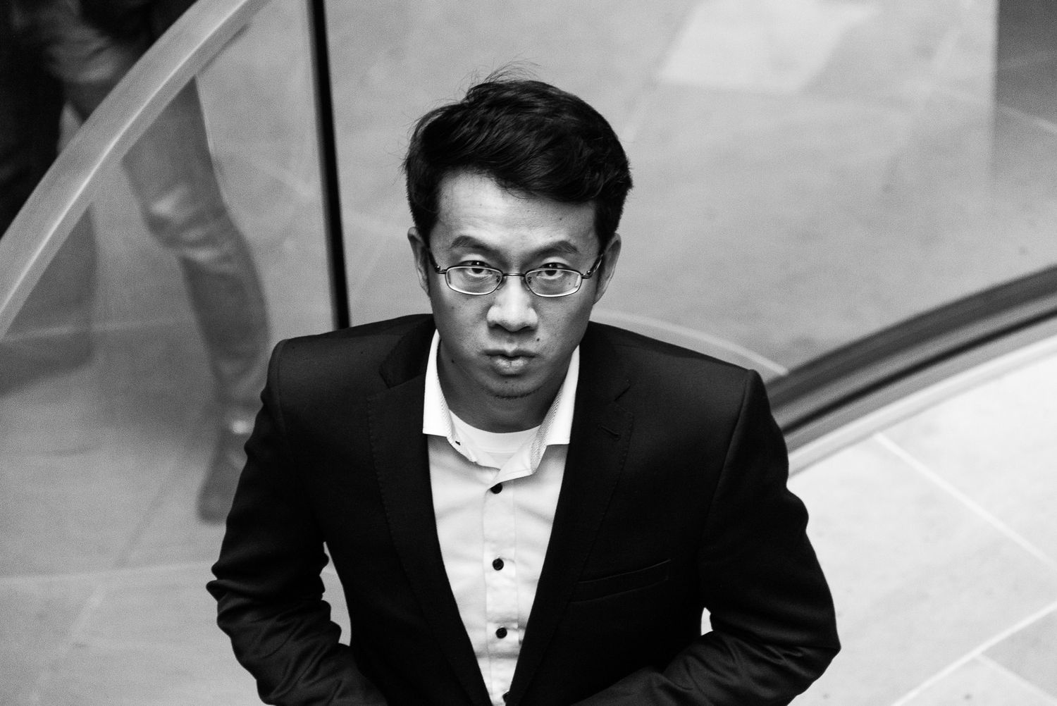 Young Asian man in tuxedo looking up at camera in black and white architectural interior photo.
