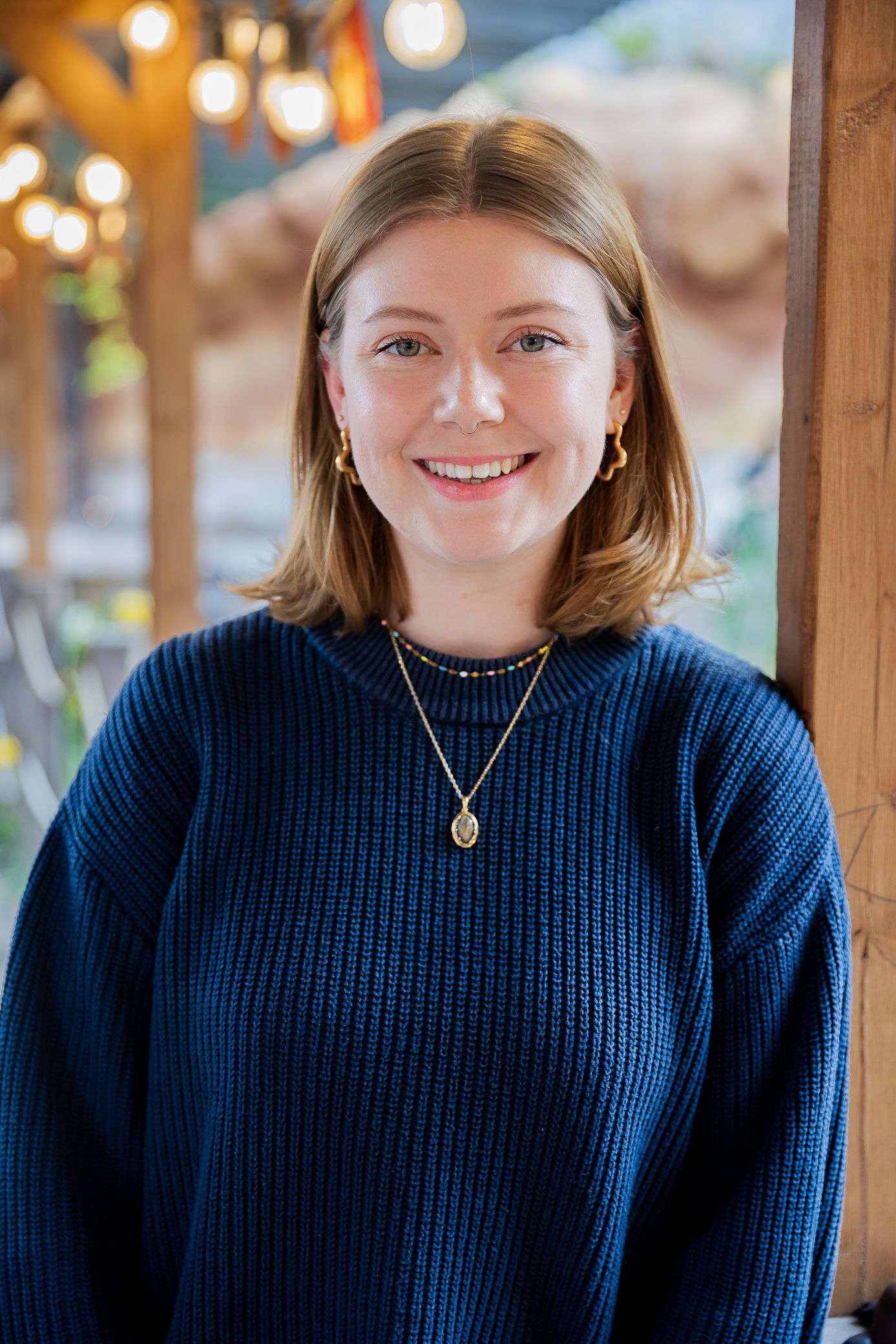 Smiling young woman with blonde bob haircut wearing a navy blue knit sweater and gold necklace indoors.