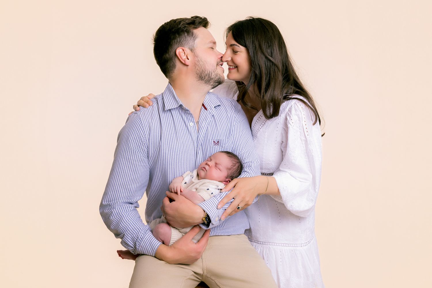 Happy couple kissing while holding their newborn baby against a warm beige background.
