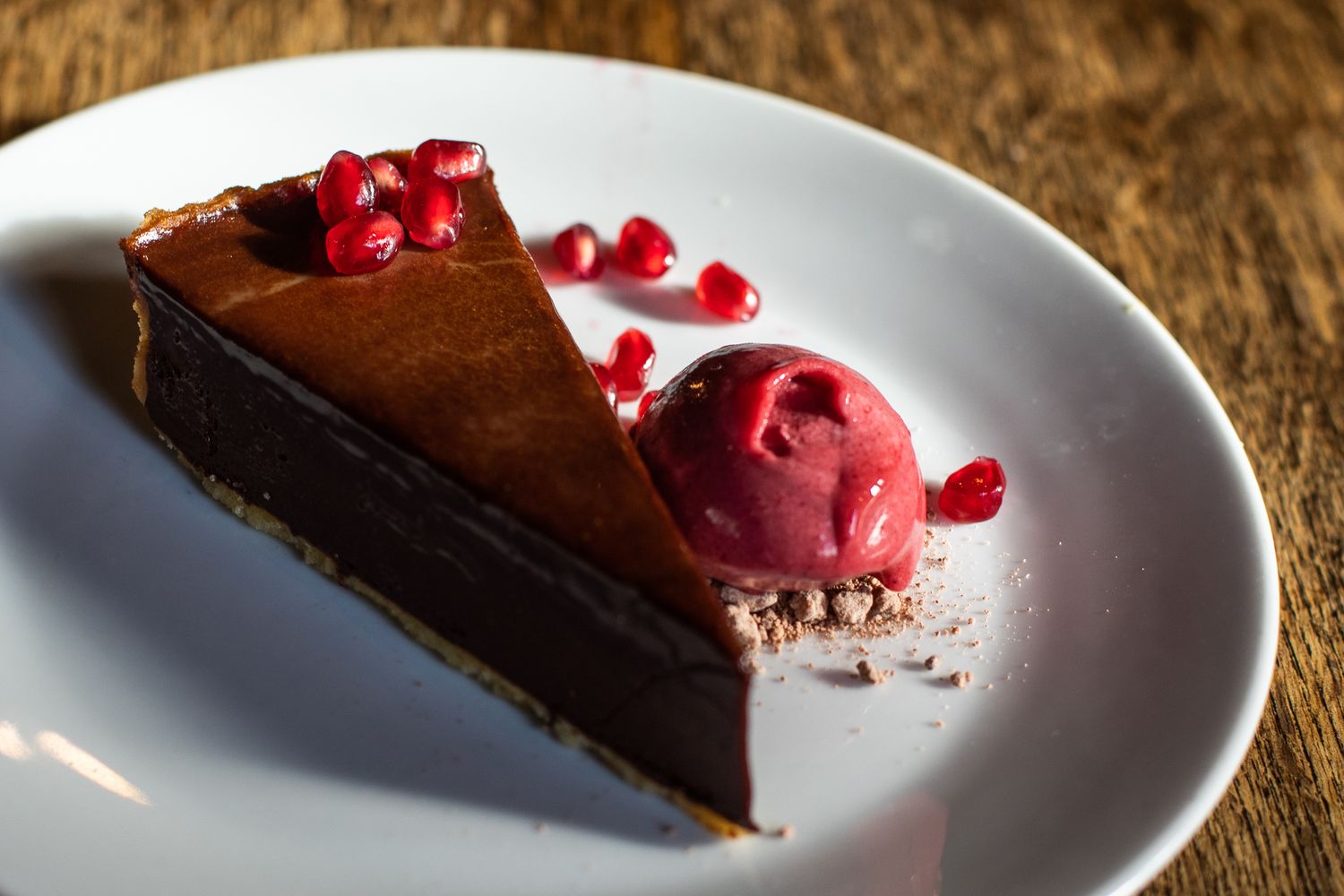 Slice of dark chocolate tart with raspberry sorbet and pomegranate seeds on white plate.
