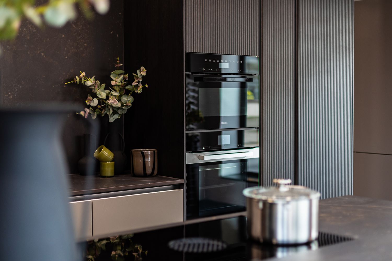 Elegant dark kitchen featuring built-in double ovens, induction hob, and stainless steel pot with floral decor.