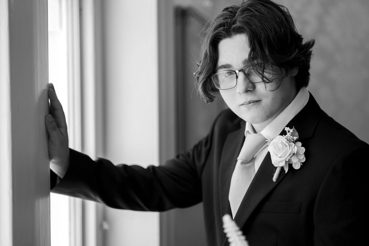 Young groom in black suit with floral boutonniere and tie poses by window in black and white wedding photo.