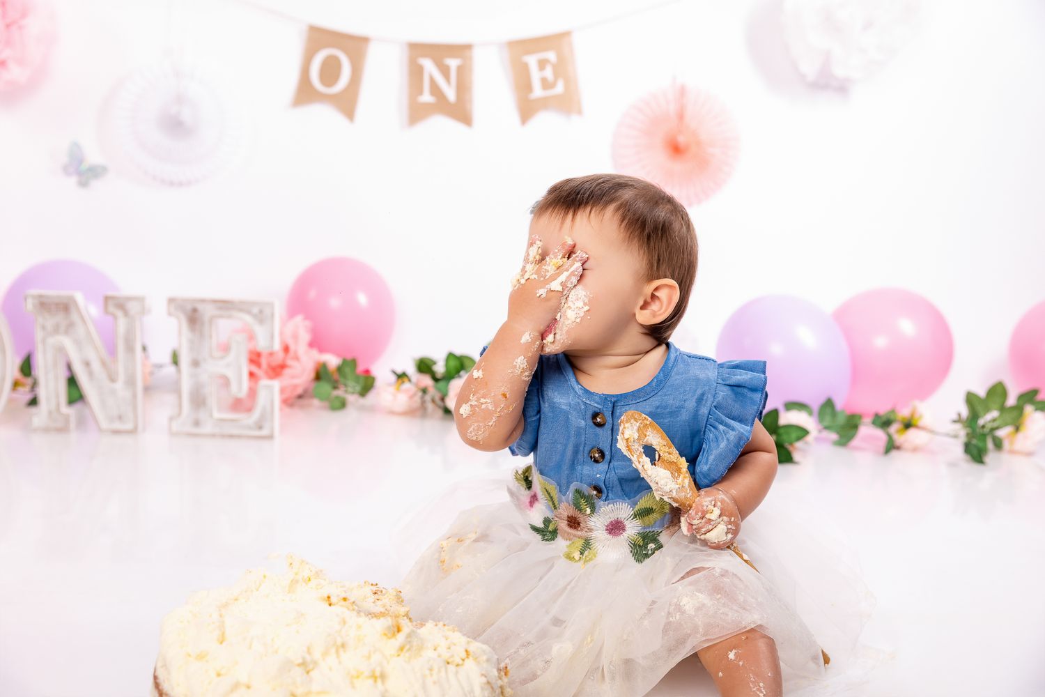 Baby girl covering face with cake during first birthday smash cake photo session with pink decor.