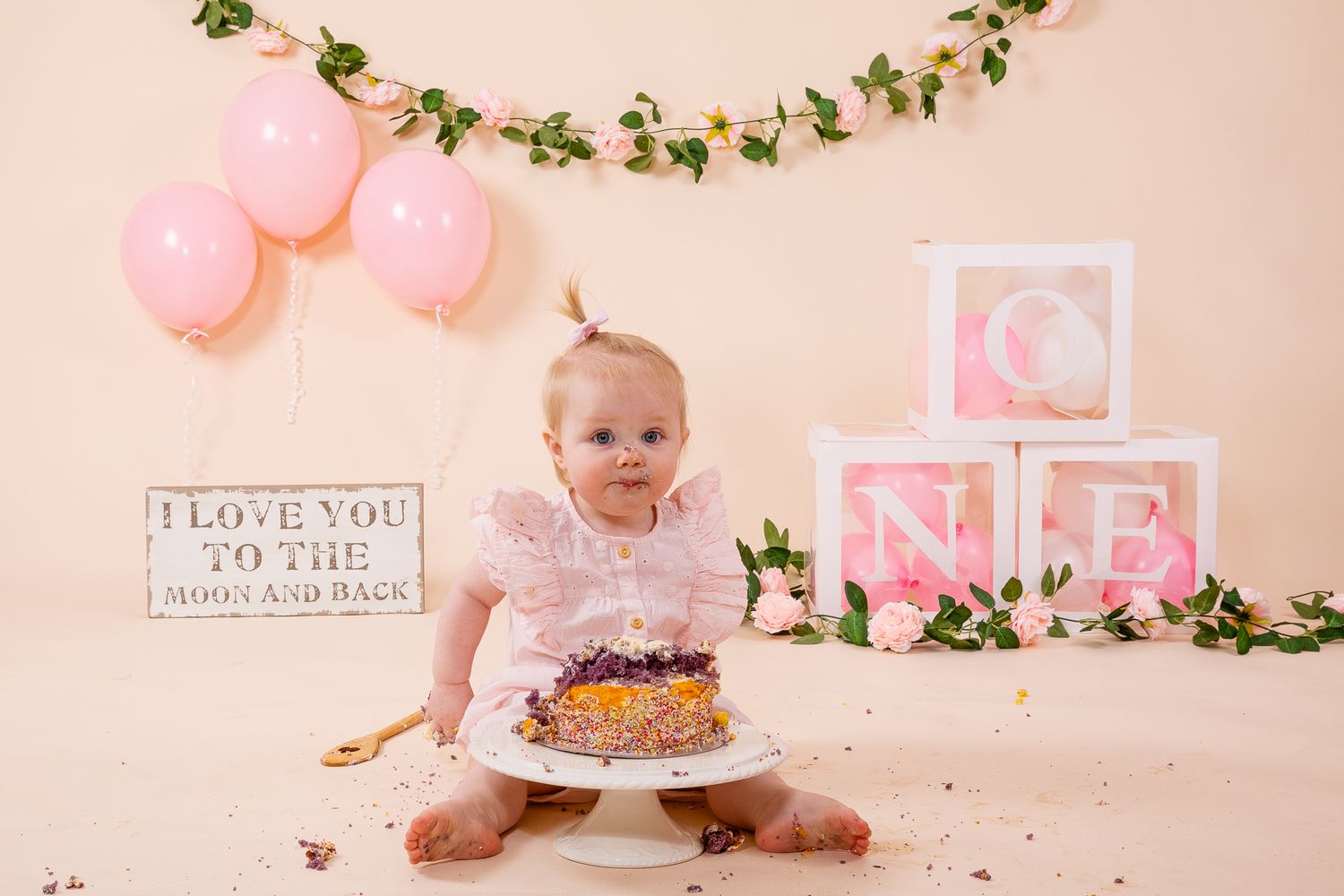 Baby girl celebrating first birthday cake smash with pink balloons, floral garland, and ONE blocks on beige backdrop.