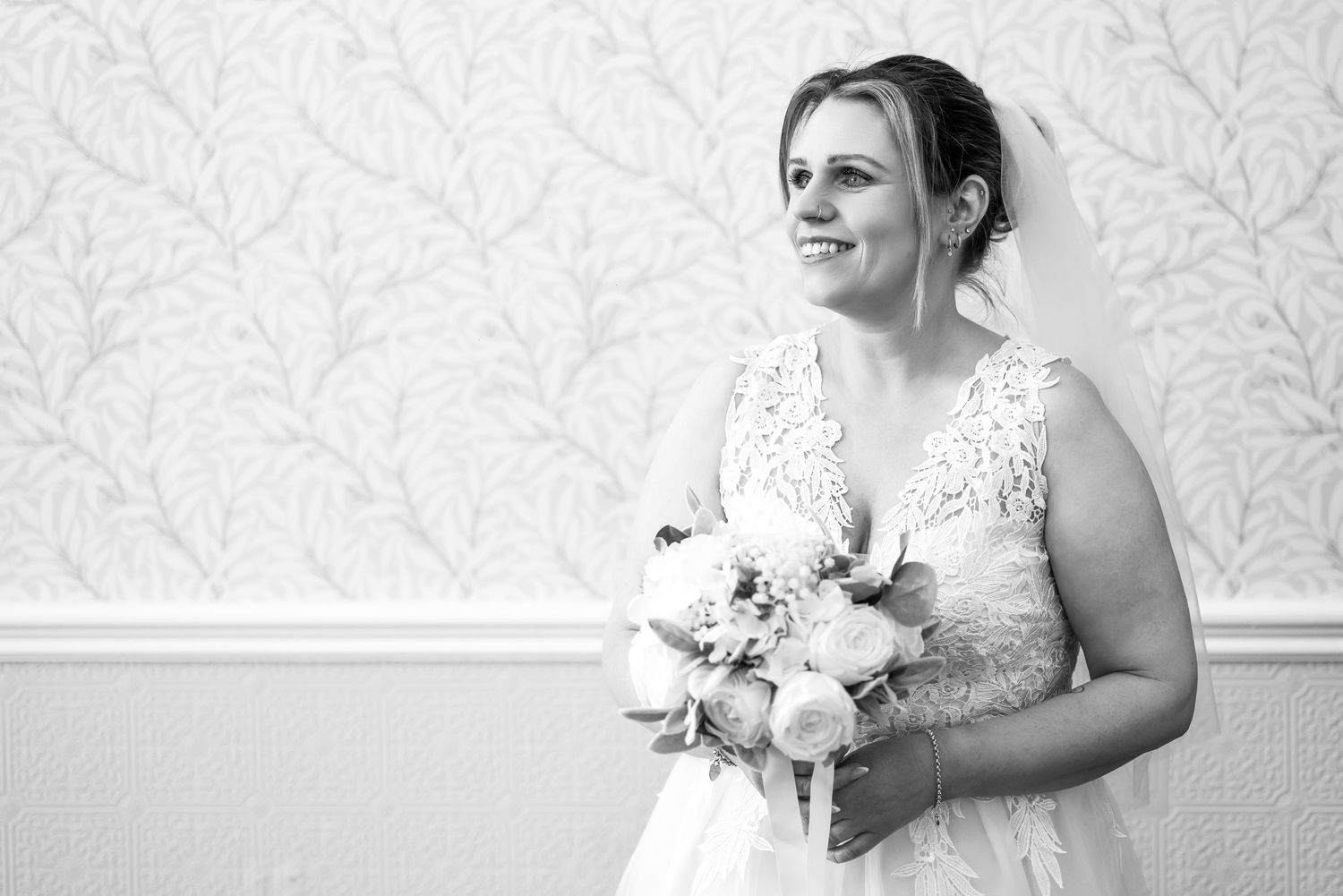 Black and white photo of a smiling bride in a lace wedding dress holding a bouquet, standing against a floral wallpaper.