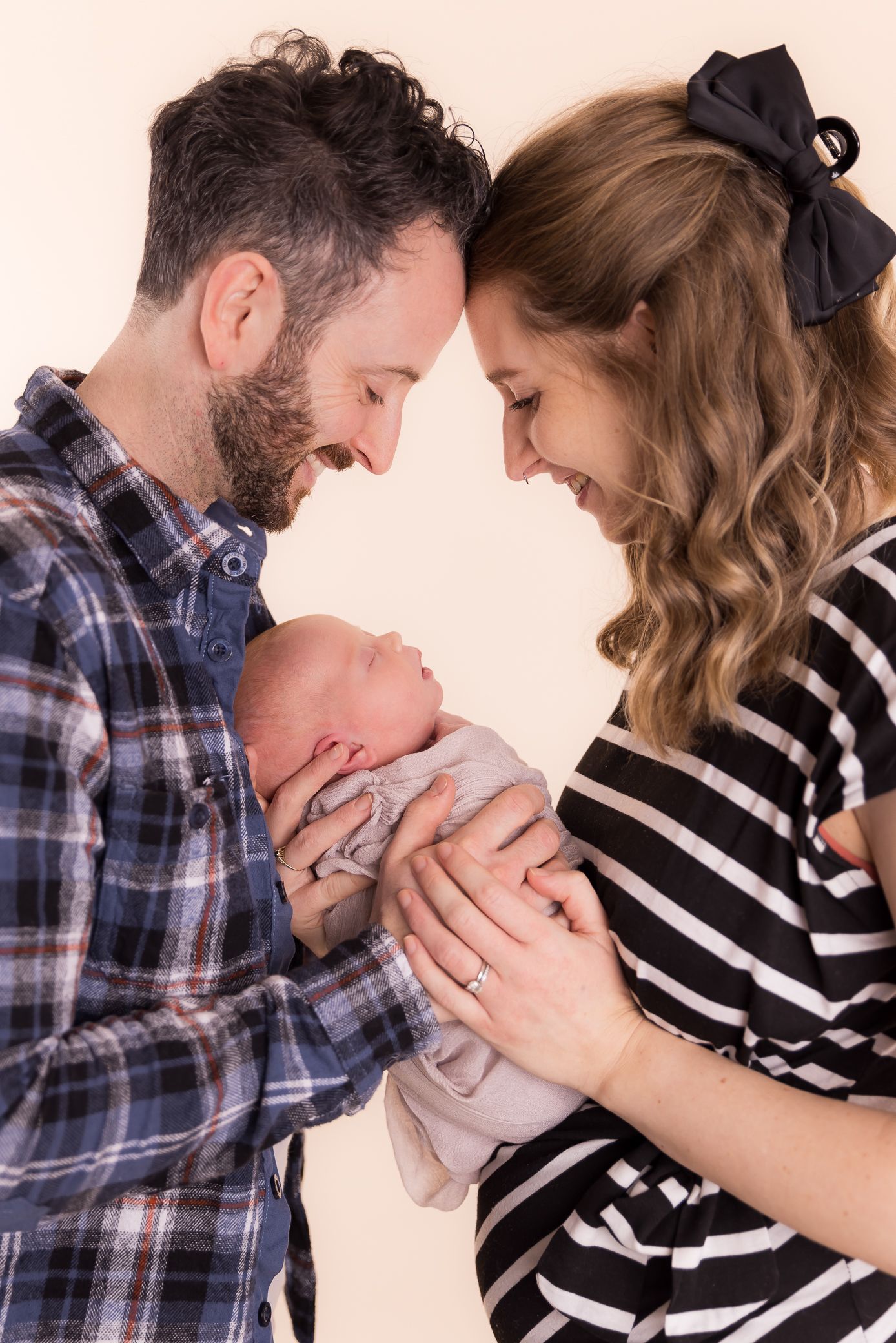 Smiling new parents touching foreheads while holding their newborn baby in a studio portrait.