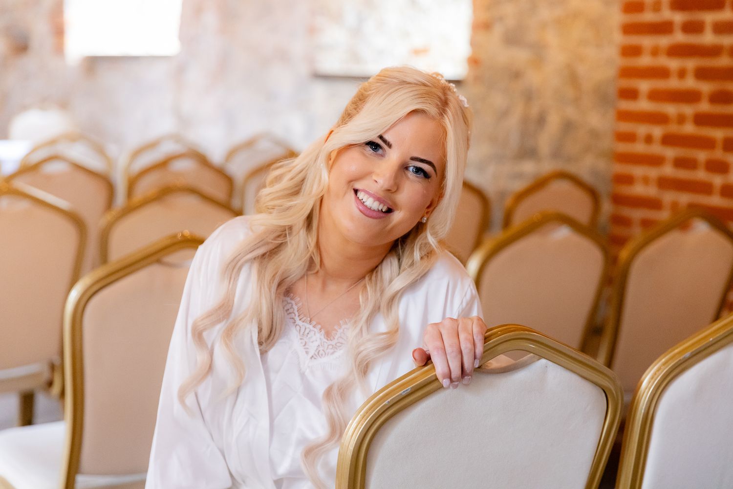 Smiling blonde bride in white robe sitting among gold chairs in elegant wedding venue.