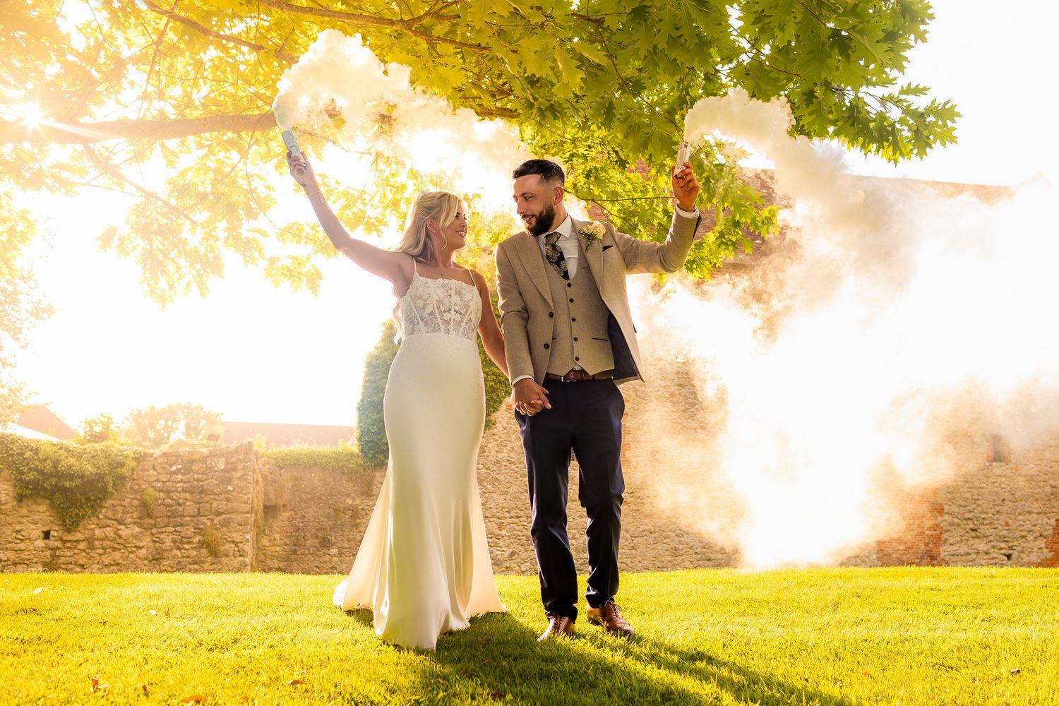 Joyful newlyweds walking through golden sunset with bouquet and colorful smoke flare.