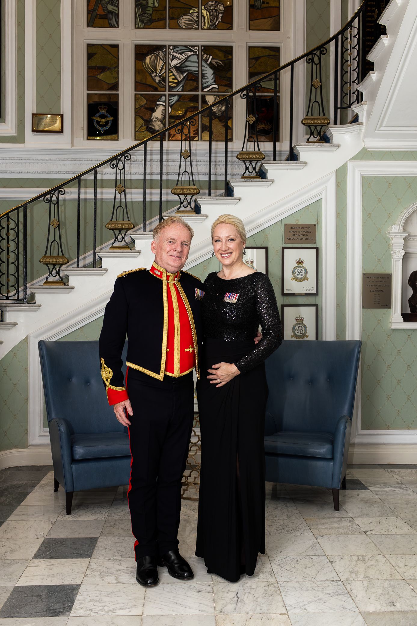 Couple posing on a marble staircase, man in red military uniform and woman in black gown.