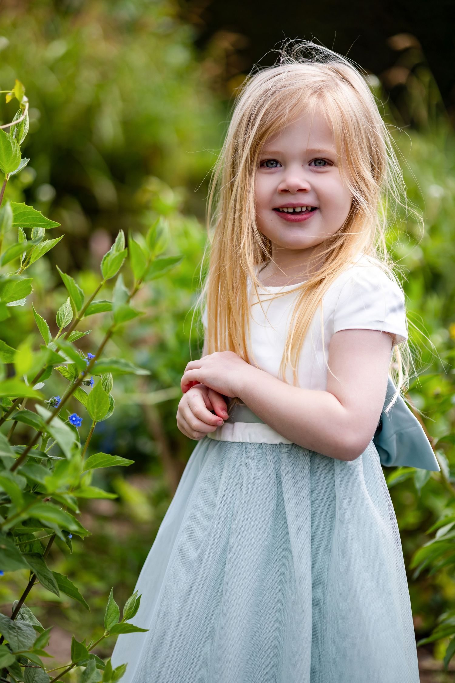Young blonde girl in a light blue tulle dress smiling outdoors among green garden plants.