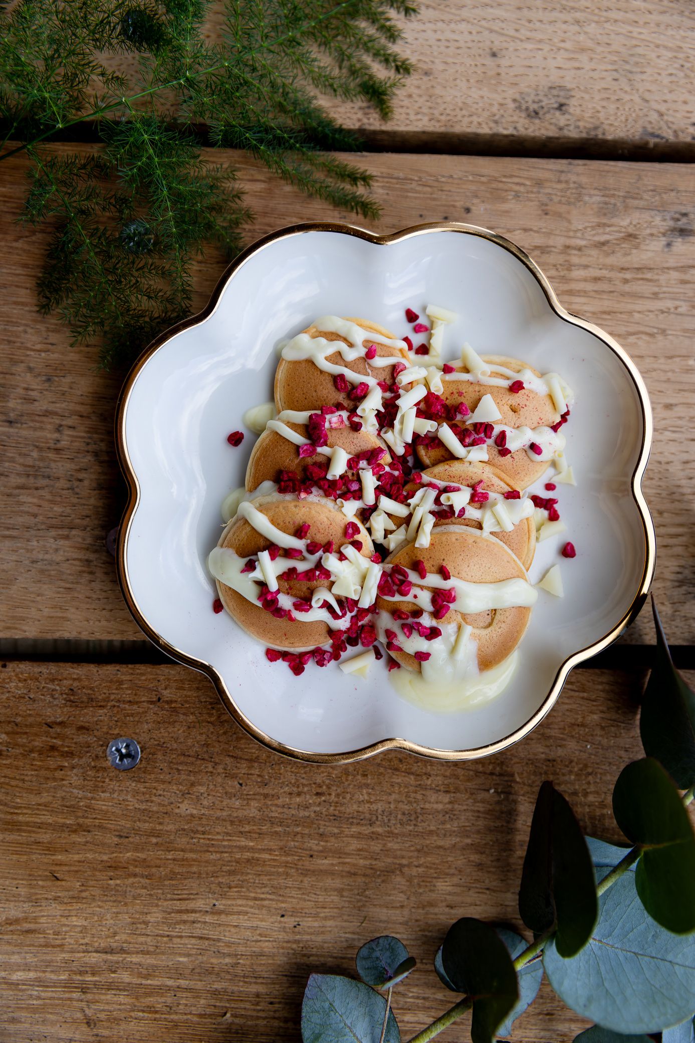 Overhead view of mini pancakes topped with white chocolate drizzle and pomegranate seeds on a plate.