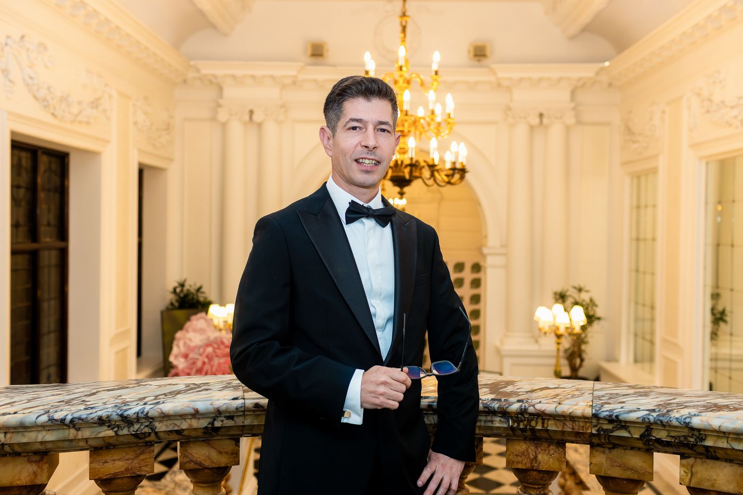 Man in black tuxedo smiles while standing at elegant marble balcony interior.