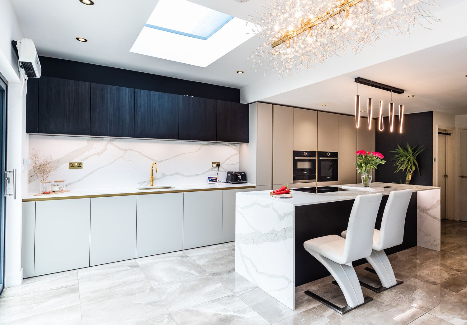 Luxury modern kitchen with marble island, dark upper cabinets, gold fixtures, and elegant chandelier.